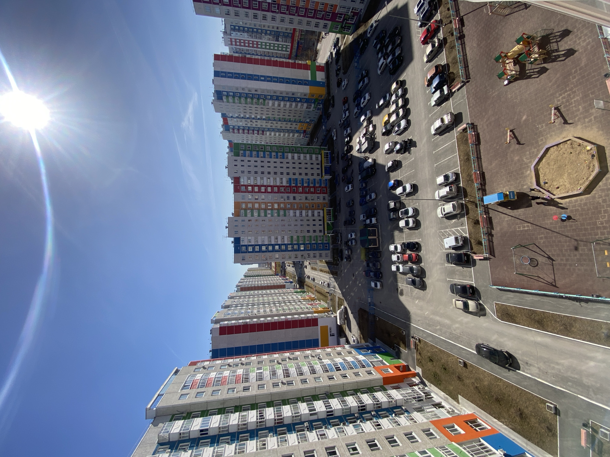 An aerial view depicts a residential district in Tyumen, Russia, characterized by tall, colorful apartment buildings. Parallel to the buildings, a large parking lot is filled with numerous cars, predominantly in shades of white, black, and grey, and some red. Adjacent to the parking area is a playground featuring a sandbox and play structures, with several small figures visible, suggesting children or adults. The environment is bright, under a clear, blue sky with the sun prominently shining. A road runs parallel to the buildings and parking lot, showing a single black car in motion.