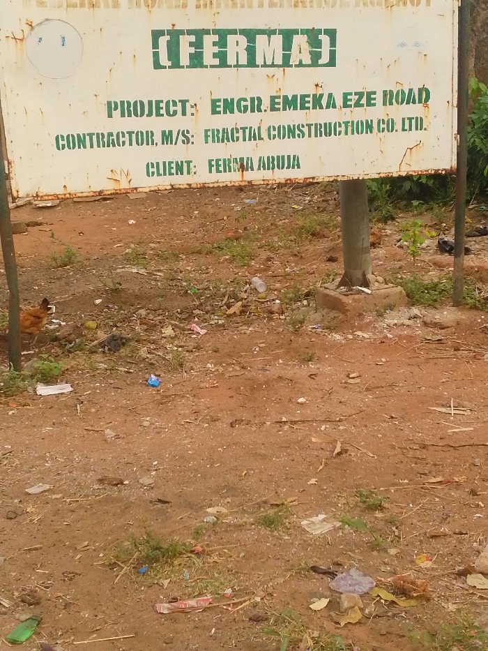 The image shows a roadside construction sign indicating a project undertaken by the Federal Roads Maintenance Agency (FERMA). The project is identified as "Engr. Emeka Eze Road," with Fractal Construction Co. Ltd. listed as the contractor. FERMA Abuja is noted as the client. The sign is mounted on a metal pole. In the foreground, the ground is covered with dirt, litter, and some sparse vegetation. A chicken is visible near the left edge of the image.