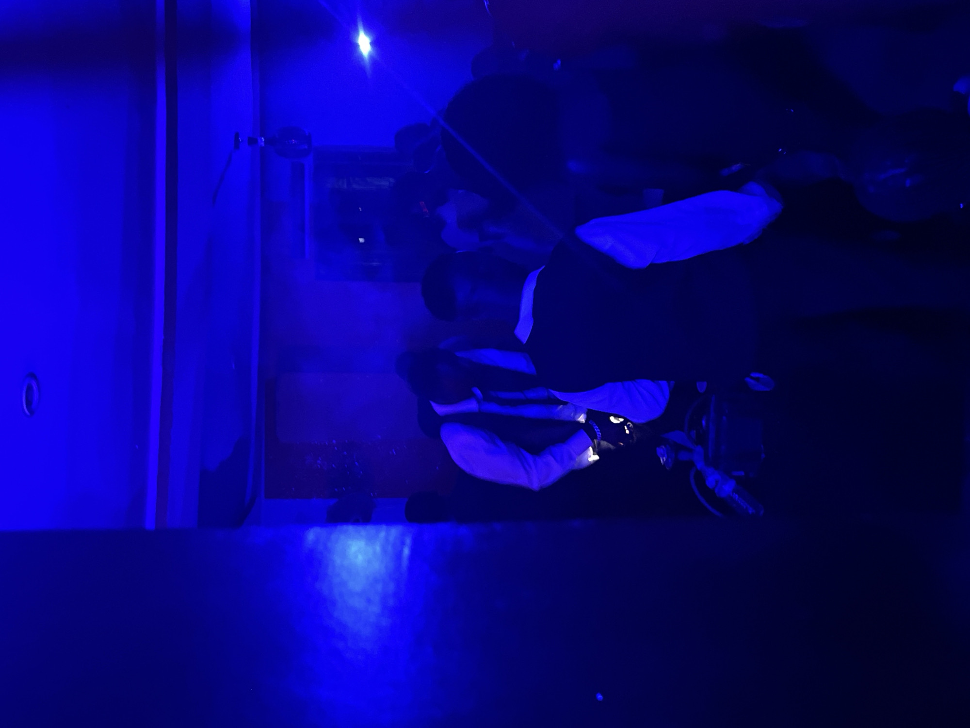 In the city of Durumi, Nigeria, a group of people are gathered in a dimly lit indoor space, illuminated by intense blue lighting. Several individuals, appearing to be young men dressed in formal attire, are visible. One person in the foreground is wearing a white shirt and a dark vest, with a dark tie. Another individual, also in a vest and white shirt, is partially obscured but seems to be bending over, possibly interacting with equipment on the floor. The environment appears to be a social gathering or event, possibly a party or celebration, with a ceiling fan visible in the upper portion of the frame. The blue lighting creates a dramatic and somewhat hazy atmosphere, with some light beams or reflections visible. The overall impression is that of a lively, perhaps festive, indoor scene.