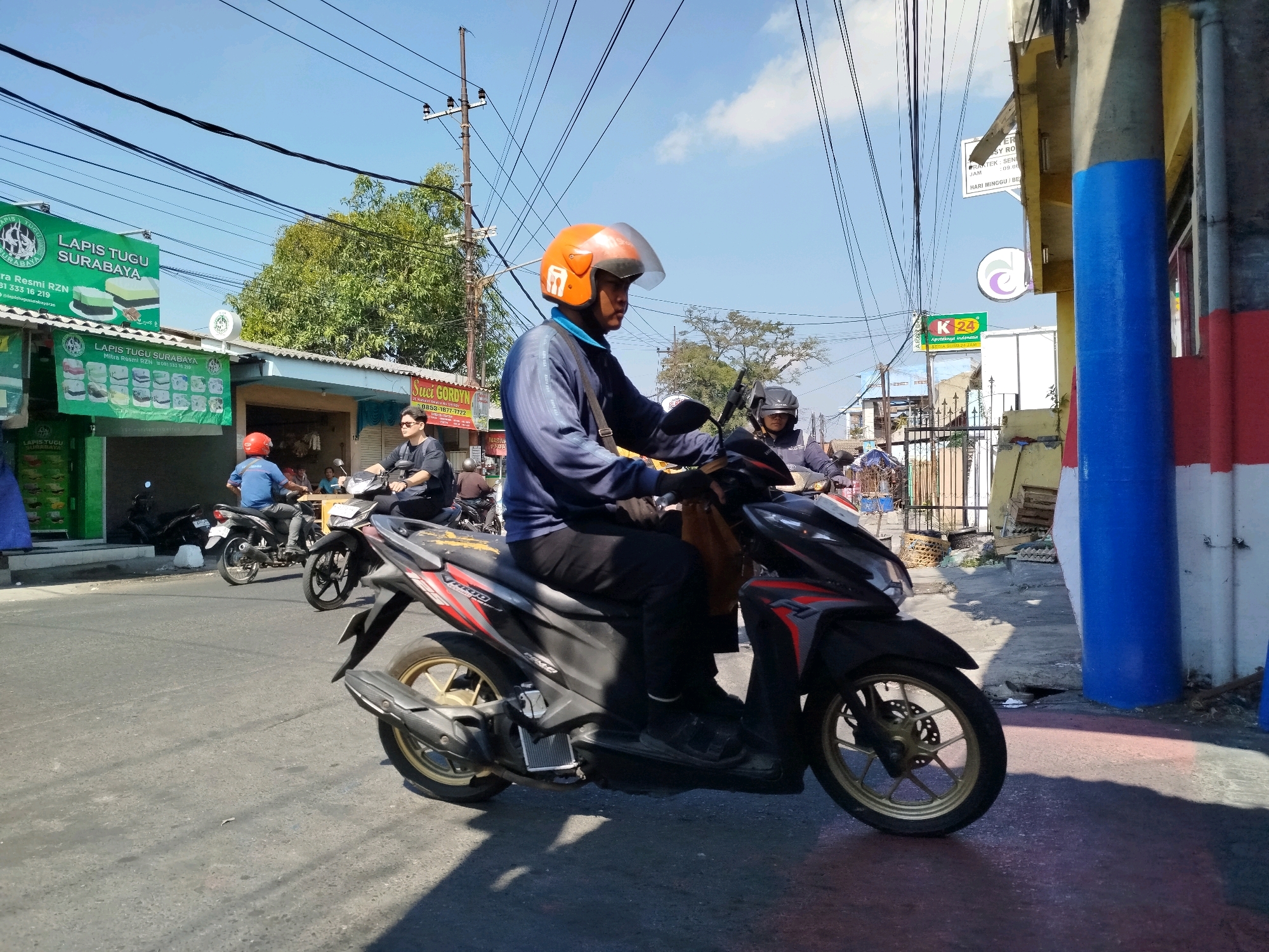 The image captures a street scene in Surabaya, Indonesia, bathed in bright sunlight under a clear blue sky with scattered clouds. In the foreground, a man wearing an orange helmet and a blue long-sleeved shirt is riding a black and red motorcycle. He is dressed in dark pants and sandals. Behind him, another motorcyclist with a black helmet is visible. The street is paved and appears to be moderately busy with several other motorcycles in the background.
Along the right side of the street, a building with a blue and white striped pillar stands prominently. Signs for "LAPIS TUGU SURABAYA" (a type of cake) are visible on the left side of the street, along with other advertisements and storefronts. The environment suggests a commercial or residential area in an urban setting. Power lines crisscross the sky, indicating a developed infrastructure. The overall impression is a typical day on a busy street in an Indonesian city.