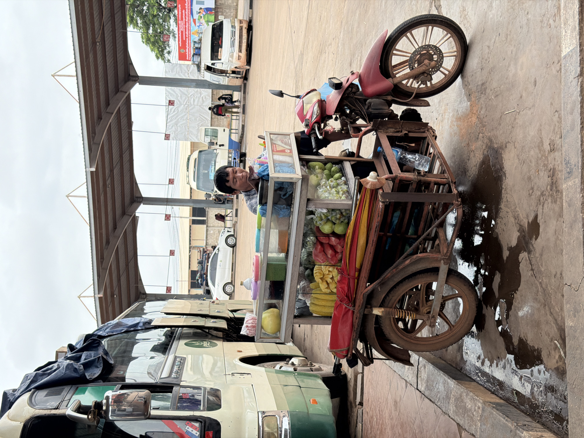 A man is sitting behind a mobile cart filled with fruits and drinks. The cart is attached to a red motorcycle. The scene is outdoors, under a covered walkway. Several buses and vans are parked in the background. The ground is wet and appears to be a bus station or transit area. The man is looking down, possibly at his products or a customer. There are no direct interactions visible between the man and other people. The environment suggests a busy transportation hub.