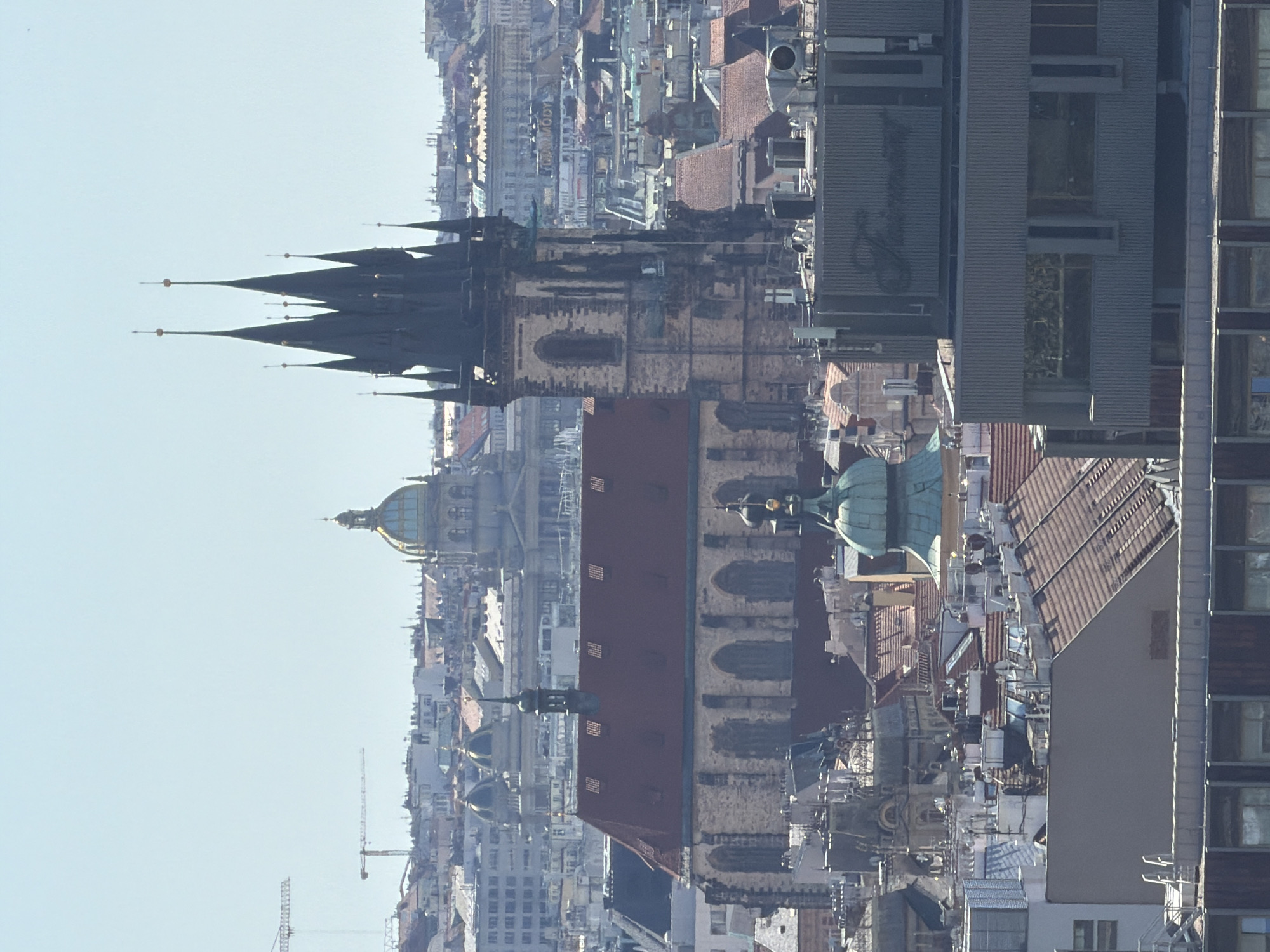 The image is a long-shot aerial view of the city of Prague, Czechia. The scene is dominated by the rooftops of buildings, with several prominent architectural features. In the foreground, a modern grey building with the word "Fairmont" visible on its facade is visible. Behind it, a church with a red tiled roof and stone walls, featuring Gothic arched windows, is prominent. To the right of the church, a dark, spired tower with multiple pointed roofs stands tall, characteristic of Gothic architecture. Further in the background, more rooftops are visible, along with several domes, one of which is golden. In the far distance, cranes suggest ongoing construction. The sky is clear and bright, indicating a sunny day. No people are visible in the image.