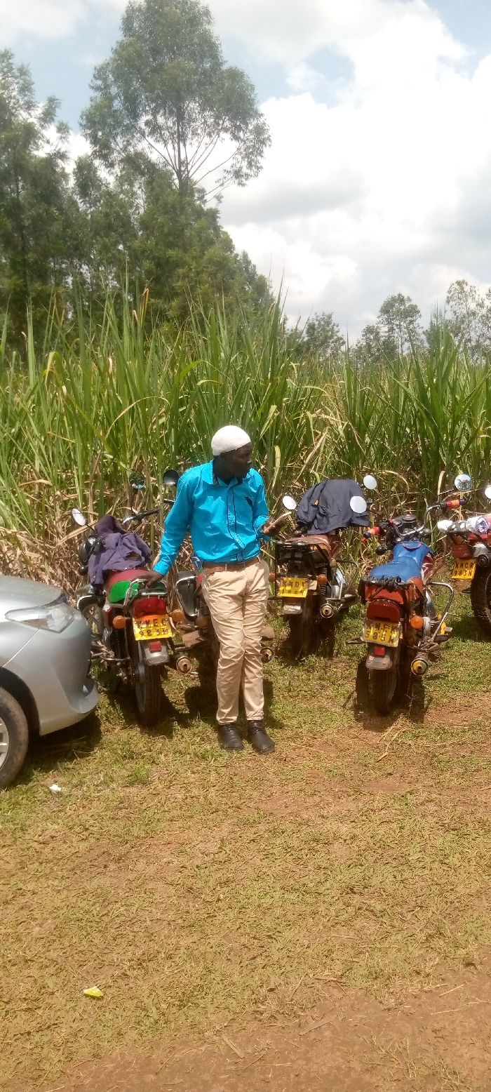 The image shows a man wearing a white cap and a light blue shirt standing in front of four motorcycles parked on a dirt road in Lwandeti, Kenya. The motorcycles have license plates visible. The man is facing the right of the frame, and his left hand is on his hip. The motorcycles are lined up from left to right. There is a light grey sedan parked behind the motorcycles. The background is a field of tall green grass. The sky is partially cloudy. The image was taken in Lwandeti, Kenya, which is located at the coordinates (latitude: 0.551, longitude: 34.801).