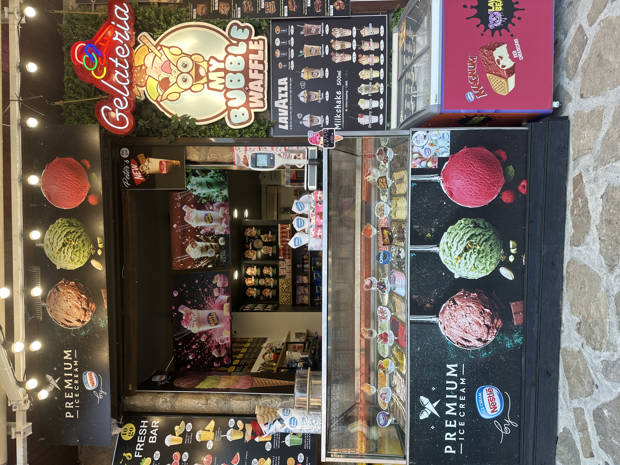 The image shows an ice cream shop called "Gelateria My Bubble Waffle" located in Nesebar, Bulgaria. The shop has a colorful facade with neon signs and large pictures of ice cream. Inside, there is a display case filled with various ice cream flavors, a coffee menu, and freezers. There are no people visible in the image, but there are advertisements for Nestle and Magnum ice cream products. The shop is situated on a cobblestone street, indicating it is likely in an older part of the city.