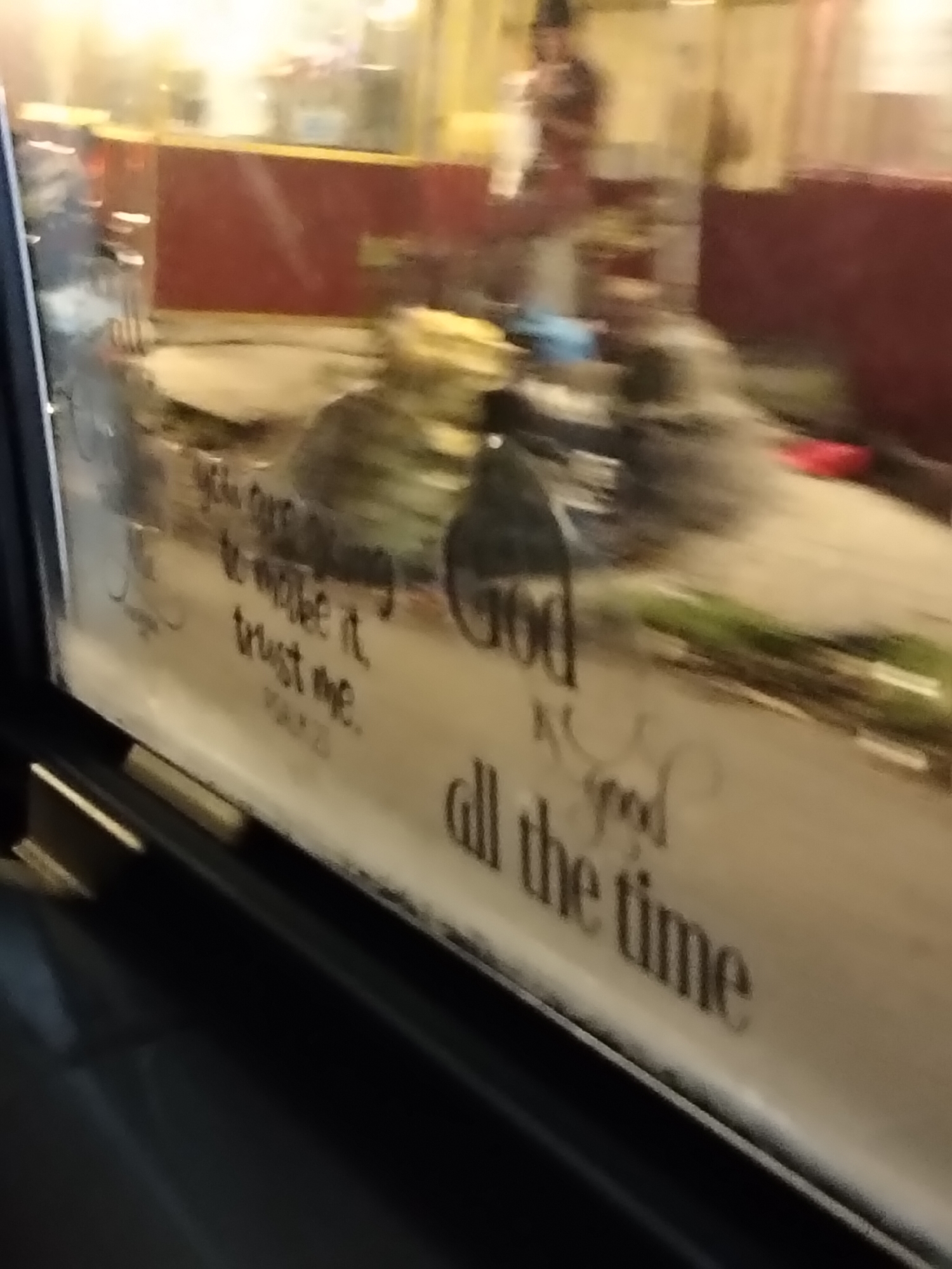 The image shows a blurry, close-up view of lettering on a window, likely from inside a vehicle. The lettering appears to be handwritten or stenciled and is in a dark color against a lighter background. The text is partially obscured by condensation or dirt on the window.
The visible text reads:
"you are
to make it
trust me.
MAX 20
God
is good
all the time"
The background is out of focus and appears to be an urban environment, possibly at night, with warm lighting and some indistinct shapes of buildings or structures. It is impossible to definitively determine the location as Davao City, Philippines based solely on the provided image.