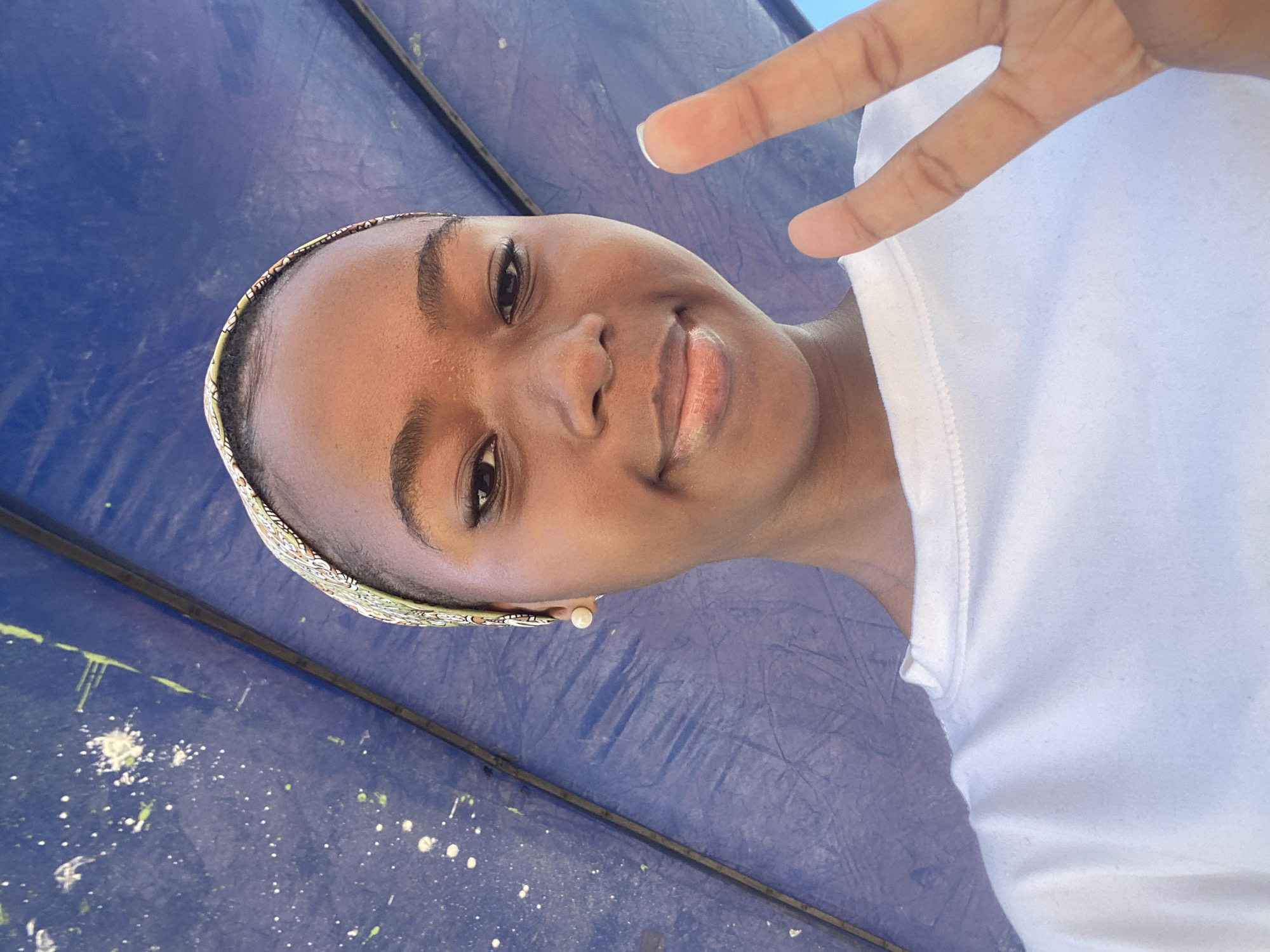 A young Black woman is seen in a close-up selfie. She wears a white t-shirt and a patterned headband. She is making a "peace" or "victory" sign with her fingers. She also wears a pearl earring. The background appears to be a blue tarp or surface with some splattered paint spots. It is not possible to determine the location in Dar es-Salaam, Tanzania from the provided image.