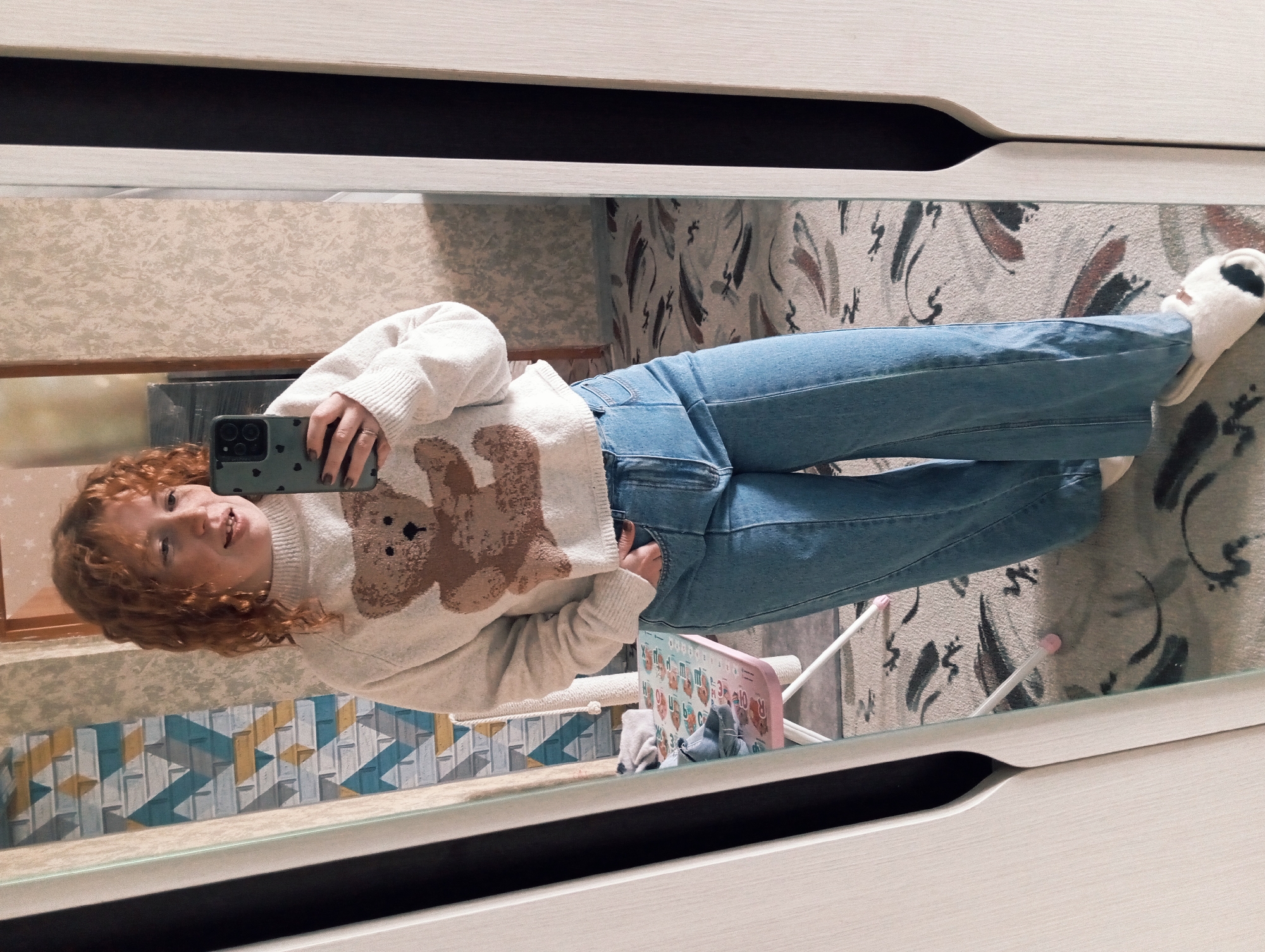 This mirror selfie captures a young individual with shoulder-length, curly red hair, reflected in a large mirror indoors. The person, appearing to be female, is dressed in a casual outfit consisting of a light cream-colored knitted sweater featuring a prominent brown teddy bear design, blue denim jeans, and white furry slippers with a black patch. She holds a smartphone with a dark green, patterned case in her left hand, positioned to take the picture, while her right hand is casually tucked into a jeans pocket. She looks towards the camera with a relaxed, friendly smile.
The background, visible through the mirror, reveals an indoor setting, likely a room in Yurgamysh, Russia. A geometric wall with blue, white, and yellow block patterns is seen to the left. On a surface below, a pink board displaying an alphabet chart with letters (e.g., "Aa Bb Cc", "Rr", "Xx") and numbers (e.g., "1 2 3") is visible, alongside part of a light-colored cat scratcher. The floor is covered by a light-toned rug or carpet featuring abstract, organic designs in shades of brown, black, and grey. The mirror itself is framed by light-colored furniture, possibly a wardrobe or dresser, with a dark handle or recess. The overall scene conveys a comfortable and casual moment.