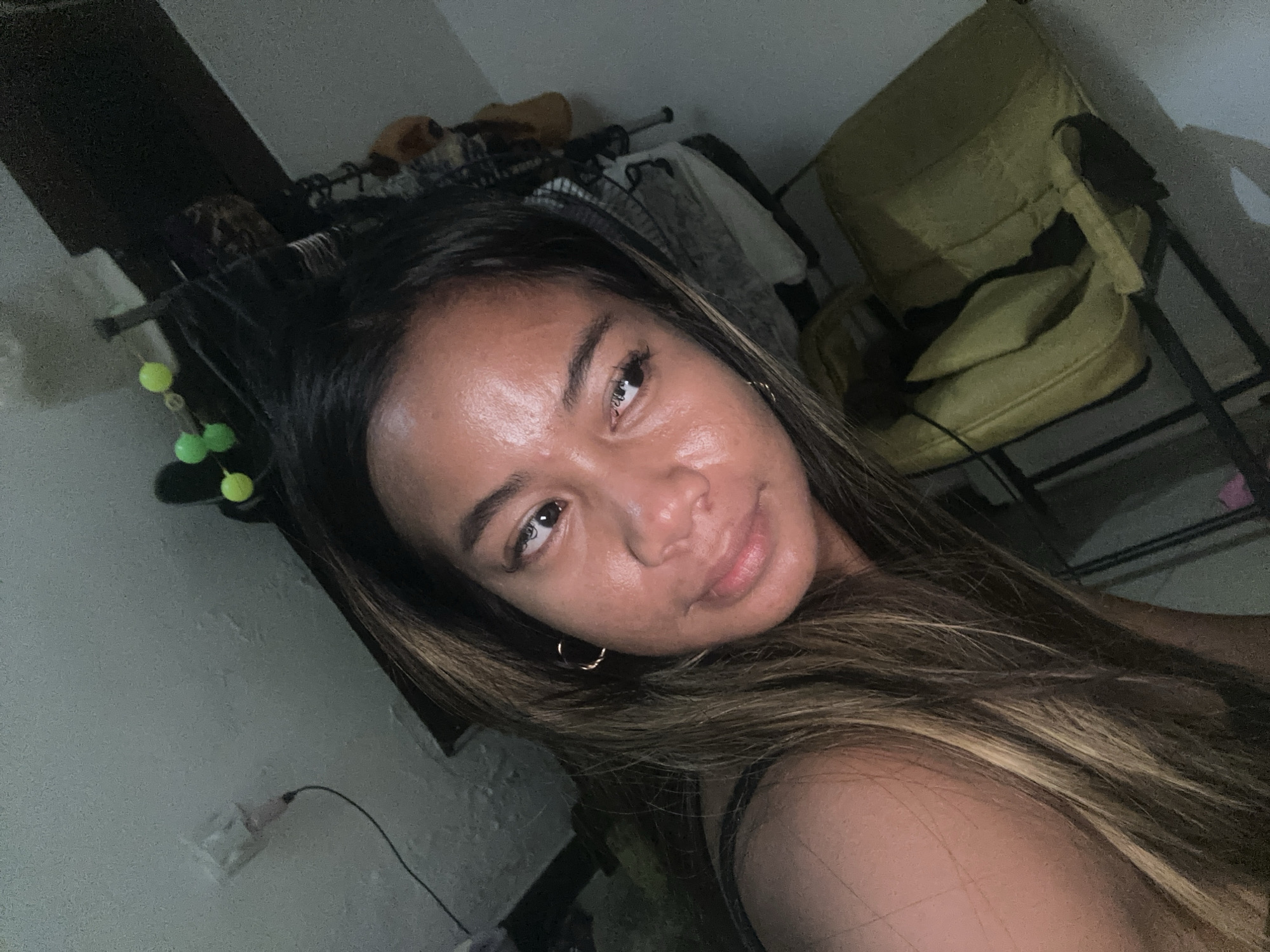 In an indoor environment, likely a residential bedroom in Sukawati, Indonesia, a young woman with long, brown hair is captured in a selfie. She is positioned with her head tilted to the left, her gaze directed towards the camera. The lighting in the room appears dim, casting shadows that obscure some details. Visible elements in the background include what seems to be a yellow armchair, a clothing rack with items of clothing, and a wall with an electrical outlet. The woman is wearing a simple dark-colored top. Her facial features, including her eyes, nose, and lips, are clearly discernible. The overall impression is that of a casual, personal moment captured in a domestic setting.