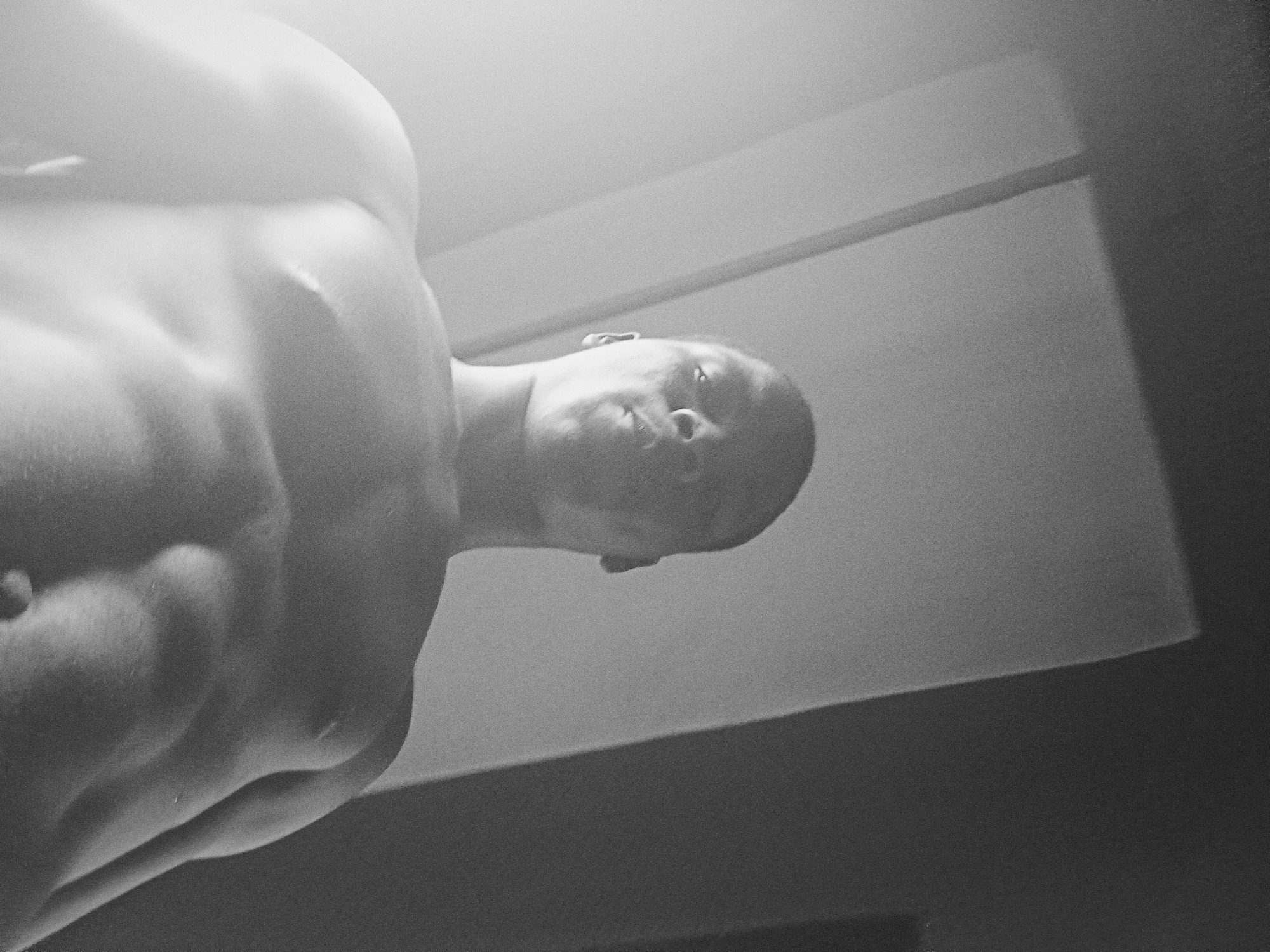 A black and white photograph shows a shirtless man with a muscular build, from a low-angle perspective. The man's head is tilted down, facing the camera. The background appears to be an indoor space with a plain wall and ceiling. The location in Nkpor, Nigeria cannot be determined from the image.