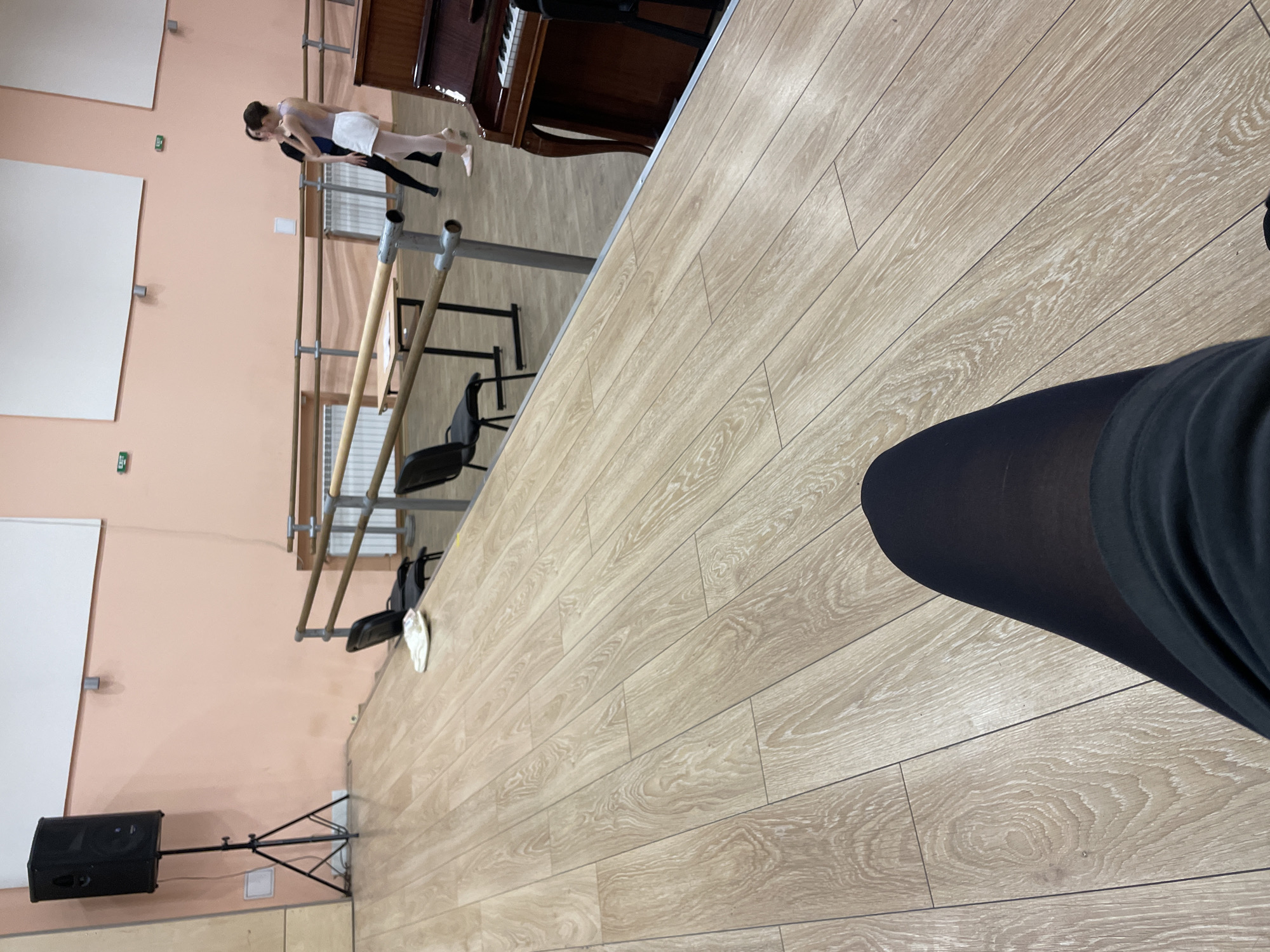 The image captures a scene within a dance studio. In the foreground, a person's legs wearing black tights are visible, resting on a polished wooden floor. The floorboards run diagonally across the frame. In the mid-ground, several black chairs are placed along a barre, which is a horizontal bar used for ballet exercises. A small table with some papers on it is positioned near the barre. Further back, two women are engaged in a dance lesson. One woman, dressed in white shorts and a purple leotard, is demonstrating a pose, while the other, in a black outfit, appears to be observing or instructing. To the right, a dark brown upright piano is partially visible. The walls are painted a pale peach color. In the background, two white rectangular panels are mounted on the wall, possibly for projections or as display boards. A black speaker on a tripod stand is located on the left side of the frame. The lighting appears to be ambient, suggesting an indoor setting. This scene is typical of a dance studio, and without further information or context relating to Plovdiv, Bulgaria, it is not possible to ascertain the specific location within the city.