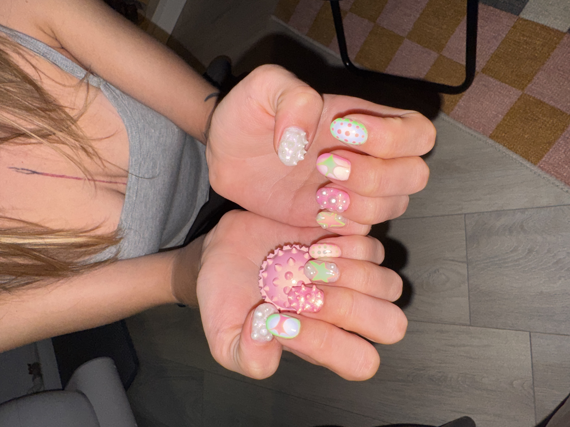The image shows a close-up of a person's hands, displaying elaborate Easter-themed nail art. The nails are adorned with pastel colors, polka dots, star shapes, and pearl-like embellishments, with some nails featuring a textured, bumpy finish. The person is holding two pink, spiky objects, one larger and one smaller, that resemble decorative Easter egg holders or toys. The hands are positioned over a wooden floor and a portion of a rug with a checkered pattern in shades of pink, brown, and grey. The background is blurred, but hints of furniture and a person's torso wearing a grey tank top are visible. The scene is indoors, but no specific location in Moscow, Russia, can be identified from the visual information provided.