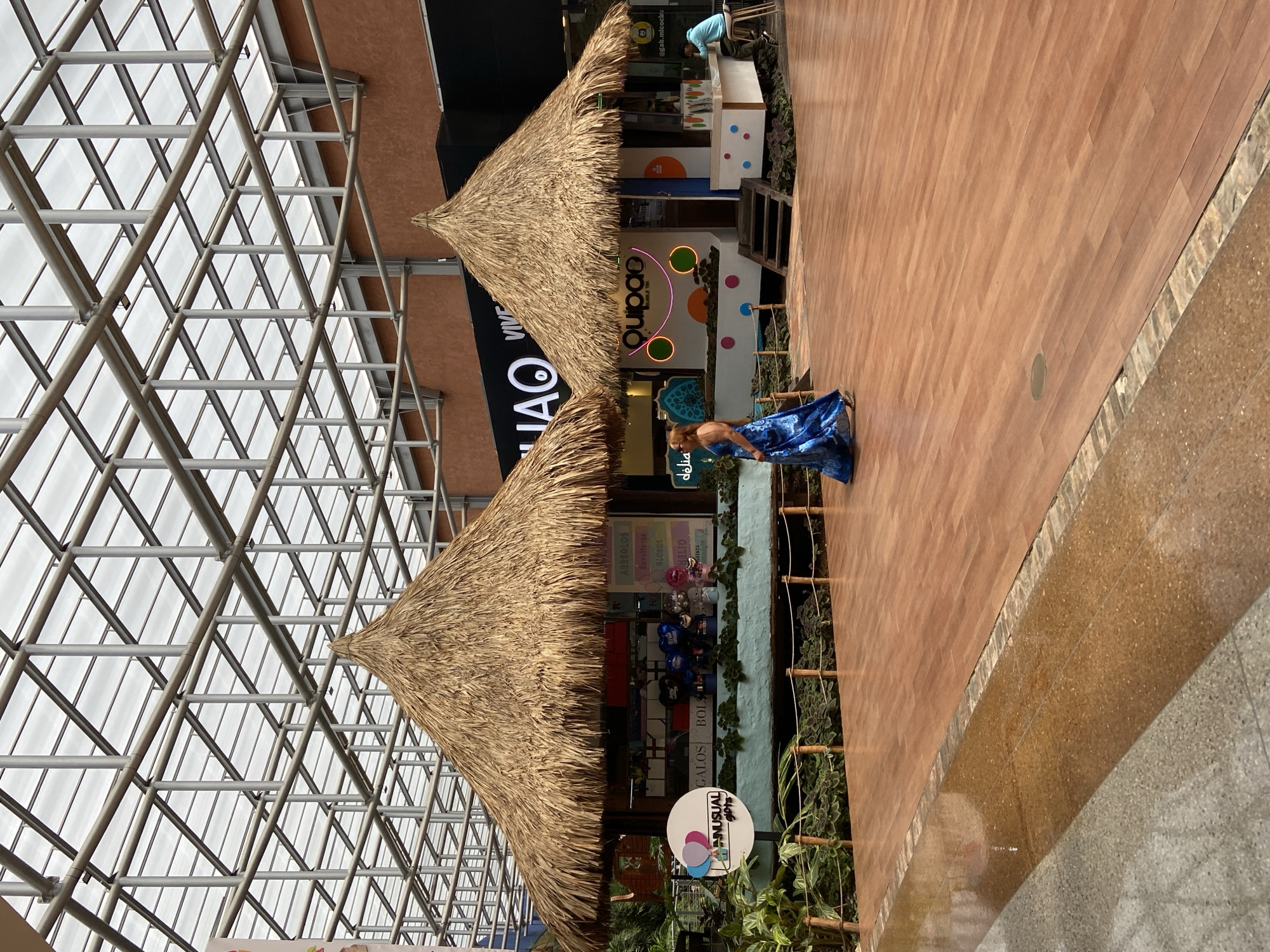 The image captures an indoor market scene in Pampatar, Venezuela, featuring two thatched-roof kiosks set against a modern, grid-like ceiling structure made of metal and translucent panels. The kiosk on the left appears to be a gift shop, indicated by signage that includes "ANUSUAL gifts," "GALOS," and "BOLS." It displays balloons and various items behind a counter. The kiosk on the right, labeled "quipao bubble tea," sells beverages. A woman in a flowing blue dress is walking between the two kiosks, her back mostly to the camera. Another person is seated at the bubble tea kiosk. The flooring is a polished wooden surface with a water feature or reflective area in the foreground. The environment is well-lit, likely from the overhead natural light filtering through the ceiling.