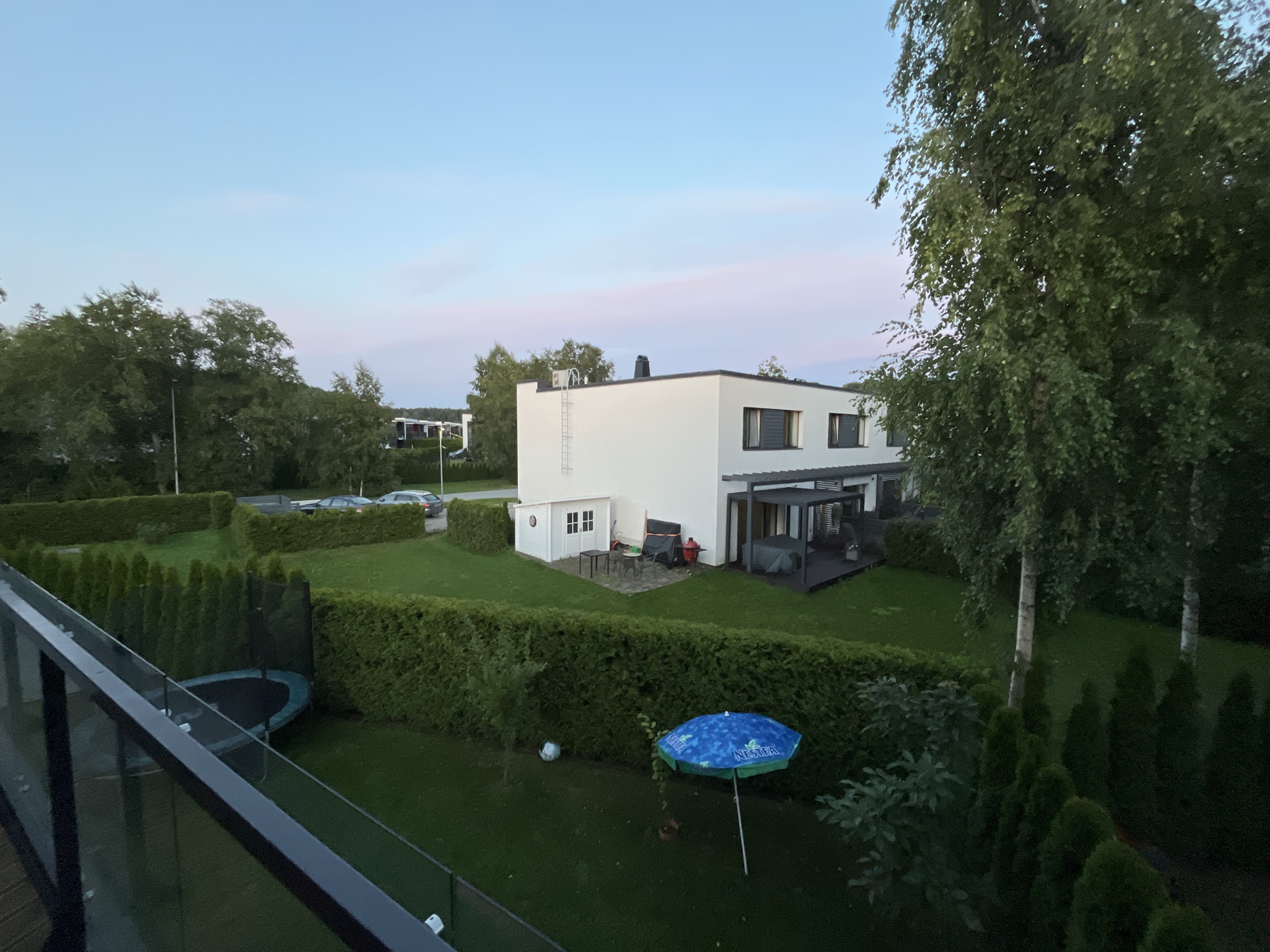 This image shows a residential yard in Haabneeme alevik, Estonia, likely taken during twilight. A modern, white, two-story house with dark-trimmed windows and a flat roof is prominent in the background. Adjacent to the house is a covered outdoor seating area with dark furniture. To the left of the house, there is a white shed with double doors and a small table and chairs outside. The yard is well-maintained with green grass, bordered by neatly trimmed hedges and evergreen trees. In the foreground, a glass-railed balcony overlooks the scene. A trampoline is visible in the left section of the yard, and a blue, patterned umbrella stands open on the grass in the middle. A soccer ball lies on the lawn near the hedges. Two cars are parked on the street in the distance, behind a line of hedges. The sky is a soft gradient of blue and pink, indicating either sunrise or sunset.