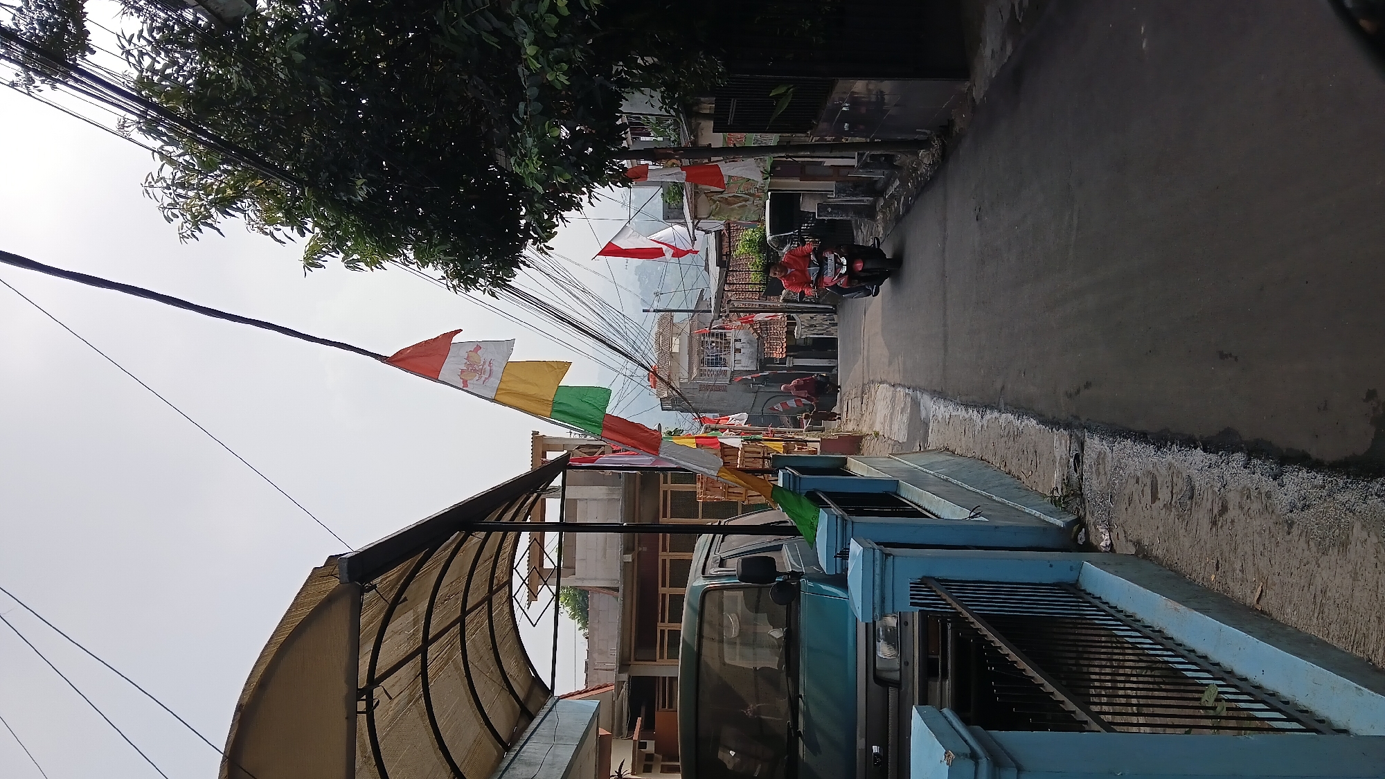 The image depicts a street scene in a densely populated residential area, likely in Bandung City, Indonesia, evidenced by the prominent display of Indonesian flags and the general architectural style. The street is narrow and paved, with a slight incline. On the left side of the street, a turquoise van is parked under a covered structure with a tan awning. A light blue fence with black metal railings runs along the edge of the property. Further down the street, a string of colorful Indonesian flags, red and white, are strung across from poles to the houses, indicating a local celebration or national holiday.
On the right side of the street, houses with red-tiled roofs are visible. A man is riding a red motorcycle towards the viewer. He is wearing a red shirt. In the distance, on the left side of the street, two figures, possibly a woman and a child, are walking away from the camera. The sky is overcast, and the overall lighting suggests it is daytime. Trees with green foliage are visible on the right side of the street, and overhead, a complex network of electrical wires crisscrosses the sky. The environment appears to be a vibrant, lived-in urban neighborhood.