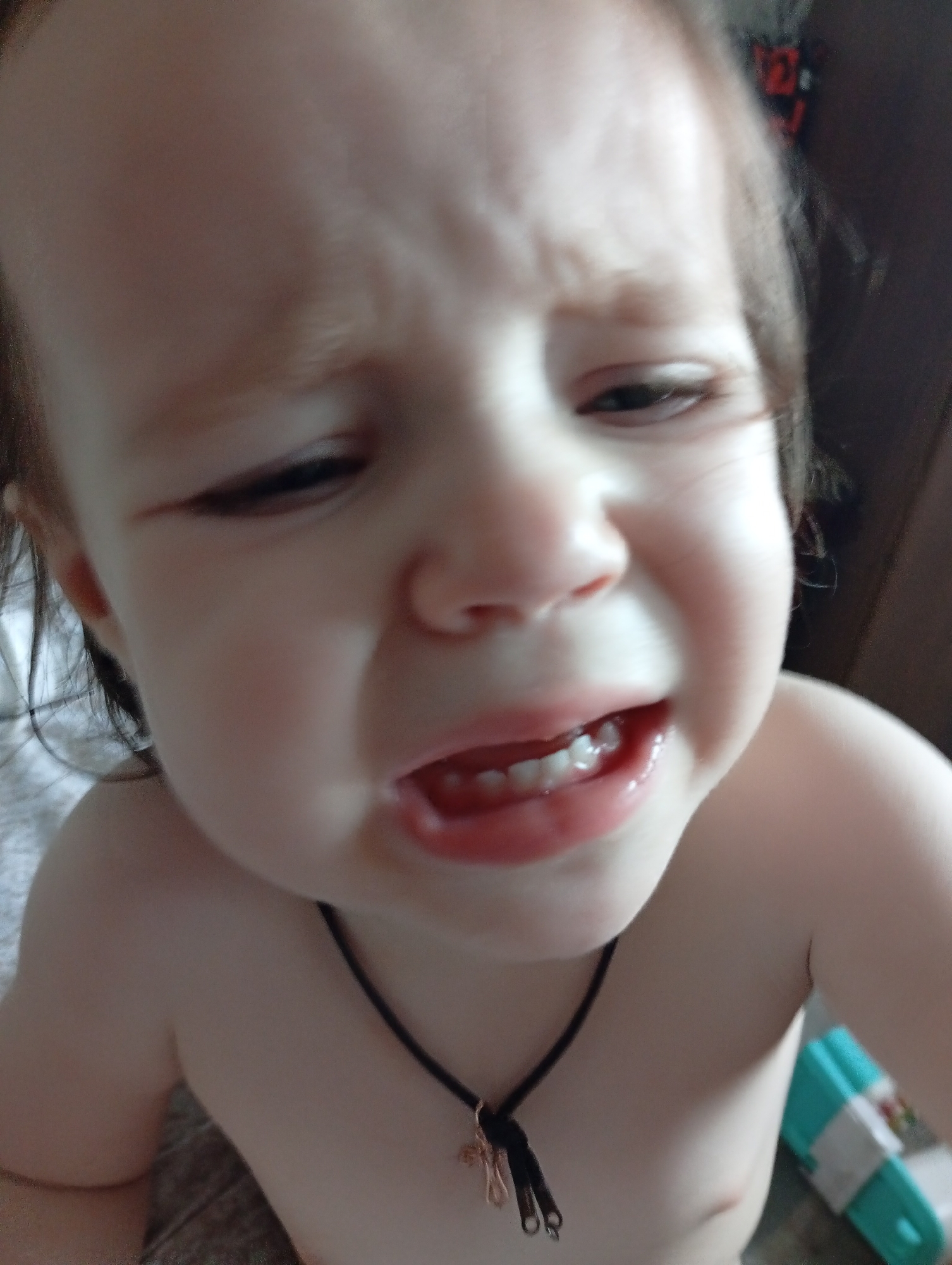 This is a close-up, over-the-shoulder shot of a young child's face, filling most of the frame. The child appears to be upset, with furrowed brows and a downturned mouth. Their eyes are partially closed, and their lips are parted, revealing several small teeth. The child is bare-chested, wearing a dark cord necklace with a small charm. The background is blurred, but suggests an indoor setting with a bed or soft surface behind the child and a darker, indistinct area to the right. The lighting appears to be artificial, given the softness and direction of the light on the child's face. The overall impression is of a moment of distress for the child.