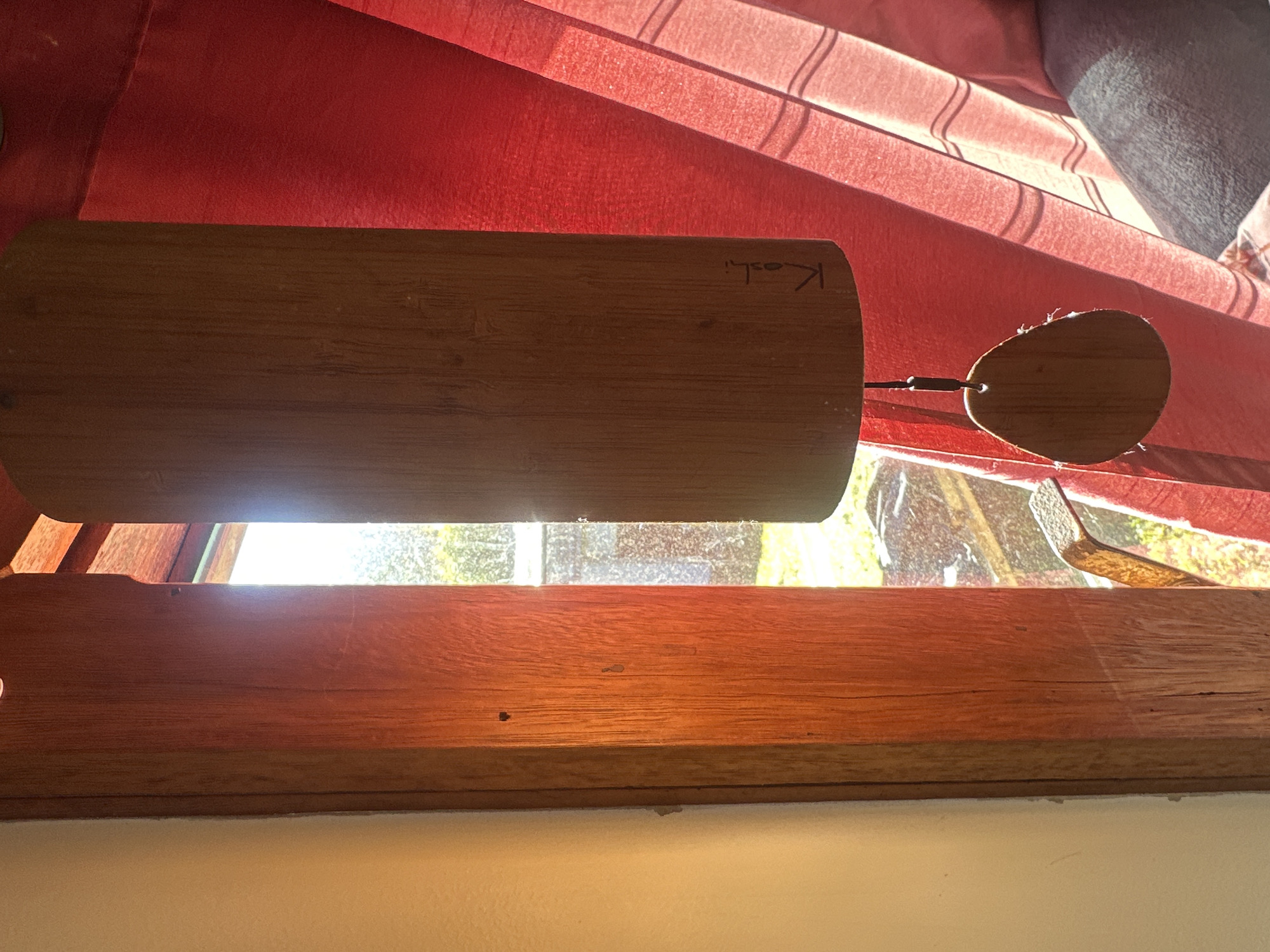 The image depicts a close-up view of a wooden wind chime hanging in front of a bright red curtain, partially obscuring a window. Sunlight streams in from the window, casting strong shadows and highlighting the textures of the wooden elements and the fabric. The wind chime appears to be made of bamboo and has a brand name "Koshi" inscribed on its surface. A smaller wooden disc hangs below the main chime. Through the window, a glimpse of the outdoors is visible, suggesting a natural or garden setting. The composition is framed by a wooden window frame on the left side, with a plain cream-colored wall to its left. There is no indication of the city of Thora, Australia, or any specific location within it. No people are present in the image, and no actions or interactions are taking place.