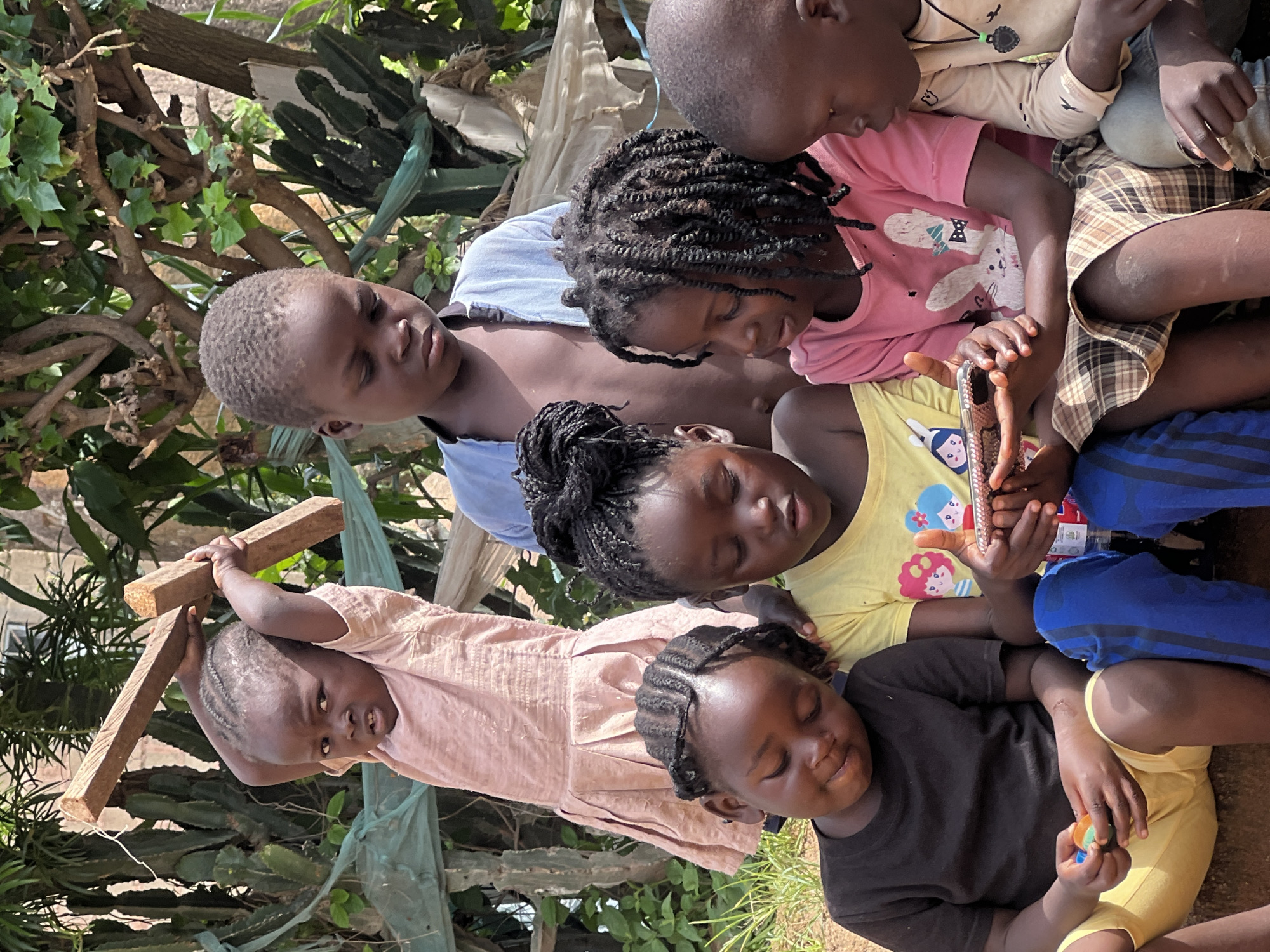 In Jos, Nigeria, a group of six children are gathered outdoors. In the foreground, four young children sit closely together. On the left, a child in a black t-shirt and yellow shorts looks down at small, colorful objects in their hands. Next to them, a child in a yellow t-shirt with cartoon characters appears to be holding a mobile phone. A third child, in a pink t-shirt with a bunny print and a plaid skirt, is also looking at the phone. To the right, a child in a beige long-sleeved shirt and jeans observes the phone. Behind them, a slightly older child in a light blue shirt stands, looking down. In the background, a very young child in a pale pink dress holds two wooden planks over their head. The children are positioned in a natural, outdoor setting with visible greenery and vegetation, suggesting a rural or semi-urban environment within Jos.
