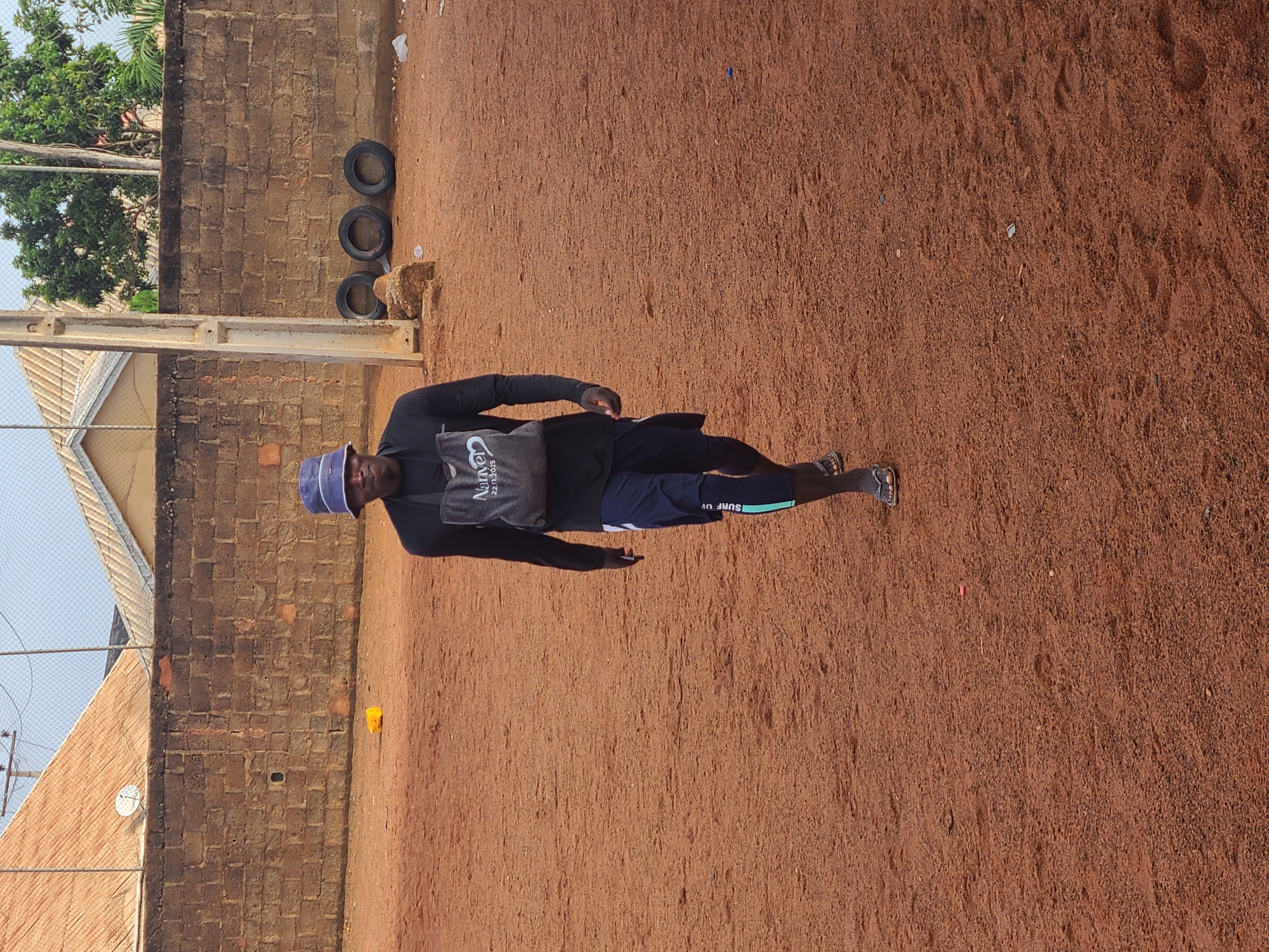 The image displays a man standing on reddish-brown earth under bright daylight. The man is positioned slightly to the left of the frame's center, facing towards the viewer with a neutral expression. He wears a dark long-sleeved shirt, dark shorts featuring a light green band with "SURF UP" printed on it, and dark sandals. On his head is a blue bucket hat. A grey sling bag with the words "Naked" and "22.11.2023" visible is worn across his chest.
The setting appears to be an outdoor, open area. Behind the man, a low wall constructed from reddish-brown bricks stretches horizontally across the mid-ground. Above and behind this wall, lush green foliage from trees is visible, along with parts of a light-colored building roof and a chain-link fence. A concrete utility pole stands vertically on the left side, close to the man. To the right of the man and against the brick wall, three dark tires are stacked. The ground, which is the dominant feature of the lower and right parts of the image, is textured with footprints and scattered small debris, consistent with laterite soil. The bright, clear lighting suggests it is daytime, likely sunny. This environment, characterized by its reddish soil, brick structures, and vegetation, aligns with the location context of Barakin Lerawi, Nigeria.