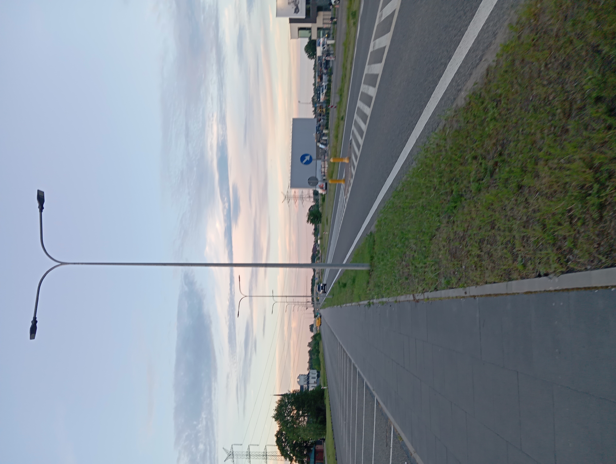 The image depicts a sidewalk next to a road in Dybów Kolonia, Poland. The sky is overcast with soft hues of blue and pink. On the left side of the image, there is a long, empty parking lot with clearly marked spaces. A sidewalk runs along the edge of the parking lot, paved with dark grey rectangular tiles. To the right of the sidewalk is a strip of green grass separating it from the road. The road itself is asphalt and has a white line marking its edge. Further to the right, there are multiple lanes of traffic and what appears to be a parking area with several vehicles. In the background, there are trees, a building that resembles a church with a tall steeple, and power lines supported by large metal towers. Several streetlights, characteristic of modern urban or suburban areas, are visible along the road. A large, rectangular billboard is present in the distance, accompanied by traffic cones and road signage. The scene appears calm and is captured during either dawn or dusk.