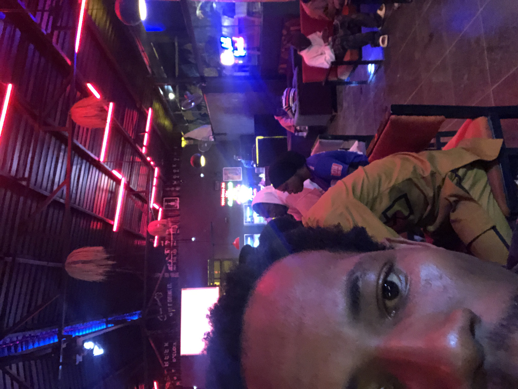 This is a medium-shot, eye-level, indoor, low-angle photograph of a group of people in what appears to be a dimly lit bar or lounge. The scene is illuminated by a combination of neon lights, primarily red and blue, that cast colored reflections on the subjects and the environment.
In the foreground, the left half of a Black male's face is prominently featured, taking up a significant portion of the image. His left eye is visible, with a dark pupil and iris, and his skin appears to be reflecting the ambient red light. His dark, curly hair is also visible at the top of his head.
In the mid-ground, other individuals are seated and standing. To the right of the foreground subject, a person wearing a yellow garment is partially visible, and behind them, another person in a blue hooded garment can be seen. Further in the background, several other people are seated on chairs and sofas, some appearing to be looking at their phones. The furniture includes dark-colored chairs and sofas with red upholstery.
The environment is characterized by a corrugated metal ceiling with numerous red neon lights running along its length. Hanging from the ceiling are decorative, spherical objects that appear to be made of straw or similar material. In the background, there are shelves with various items and what looks like a bar area with a television screen or digital display. The floor is dark and reflective, further contributing to the low-light atmosphere.
The overall impression is that of a lively but dimly lit social gathering space, possibly a bar or entertainment venue, with patrons enjoying themselves under the vibrant neon lighting. The location is stated to be Rukuba Road, Nigeria.