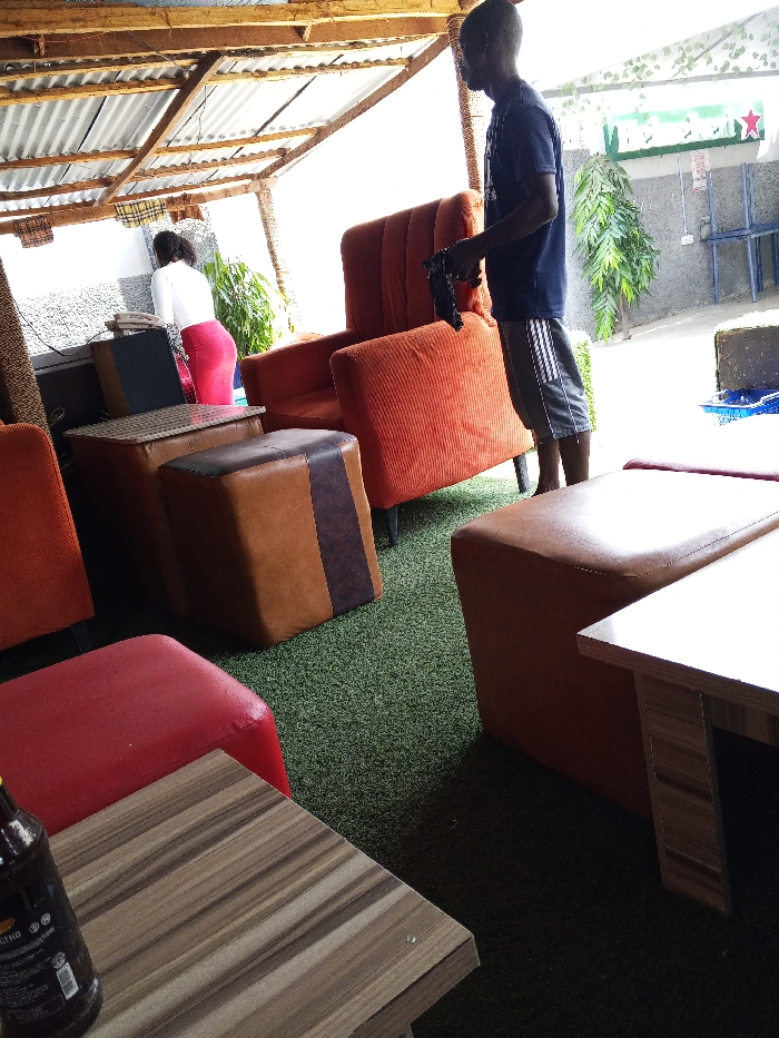 This image captures a semi-outdoor lounge area in the city of Adonkolo, Nigeria. The scene features a corrugated metal roof supported by wooden beams and a green artificial turf floor. Two individuals are present: a male standing in profile on the right, wearing a dark t-shirt and striped shorts, holding a dark object in his hands; and a female in the background on the left, facing away from the camera, dressed in a white top and red skirt, positioned near a light-colored counter. The area is furnished with multiple orange upholstered armchairs, brown and red upholstered ottomans, and small wooden tables. A dark bottle, possibly a stout, is visible on a table in the foreground. In the background, a green leafy plant and a sign with green lettering and a red star are discernible. The scene is brightly illuminated by natural light.