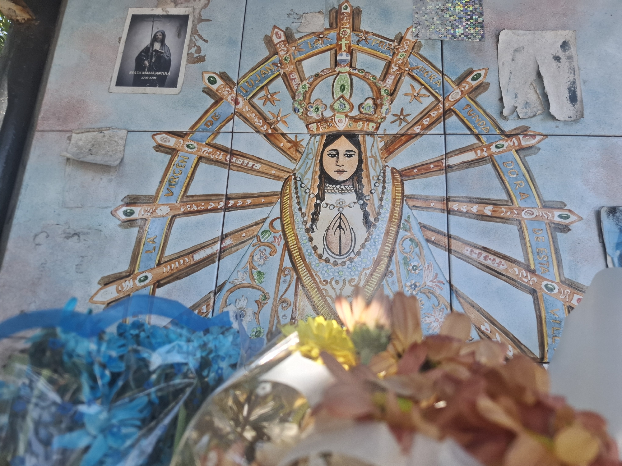 The image captures a religious shrine in Villa Domínico, Argentina. The backdrop is a tile mural depicting the Virgin of Luján, adorned with a crown and radiating beams, labeled with the words "VIRGEN DE LUJÁN," "MERCED," "FUNDADORA," and "DE ESTA VILLA." To the left, a smaller framed picture shows "BEATA MAMA ANTULA, 1730-1799." Various offerings, including blue and brown/yellow flowers wrapped in plastic, are placed in front of the mural. A patch of holographic material and a tattered white paper are adhered to the top right side of the mural. A concrete slab rests to the left of the Virgin's image. The scene suggests a place of reverence and devotion.