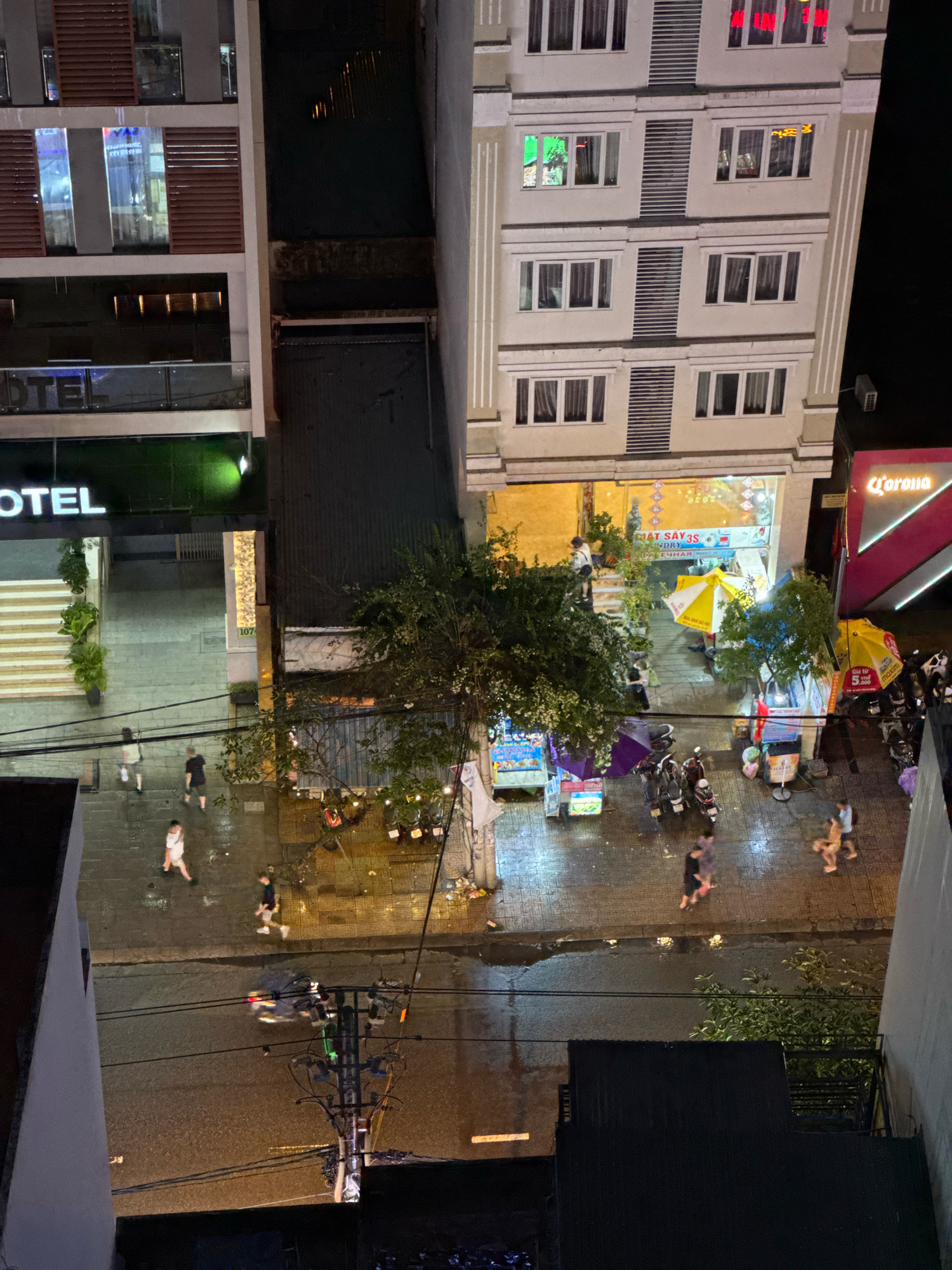 This is a night-time aerial view of a street scene in Nha Trang, Vietnam. The image captures a narrow street lined with multi-story buildings. On the left, a building with a dark facade and illuminated windows displays the word "HOTEL" in large white letters. Stairs lead up to the hotel entrance, flanked by potted plants.
The central focus is a building with a cream-colored facade, featuring multiple rows of windows. A storefront at ground level is open, revealing interior lighting and some signage, including what appears to be laundry services. Yellow and white umbrellas are set up outside this establishment.
To the right of the central building, a vibrant pink and white sign for "Corona" is visible, suggesting a bar or entertainment venue. A row of motorcycles is parked along the sidewalk. Several people are walking on the wet pavement, which reflects the streetlights and building lights. The street itself appears to have been recently rained on, with visible puddles and reflections.
Further down the street, more parked motorcycles and street vendors with carts are present. Overhead, electrical wires and a utility pole with multiple insulators are visible against the dark sky. The overall scene depicts a typical urban street at night, bustling with activity and illuminated by artificial light.
