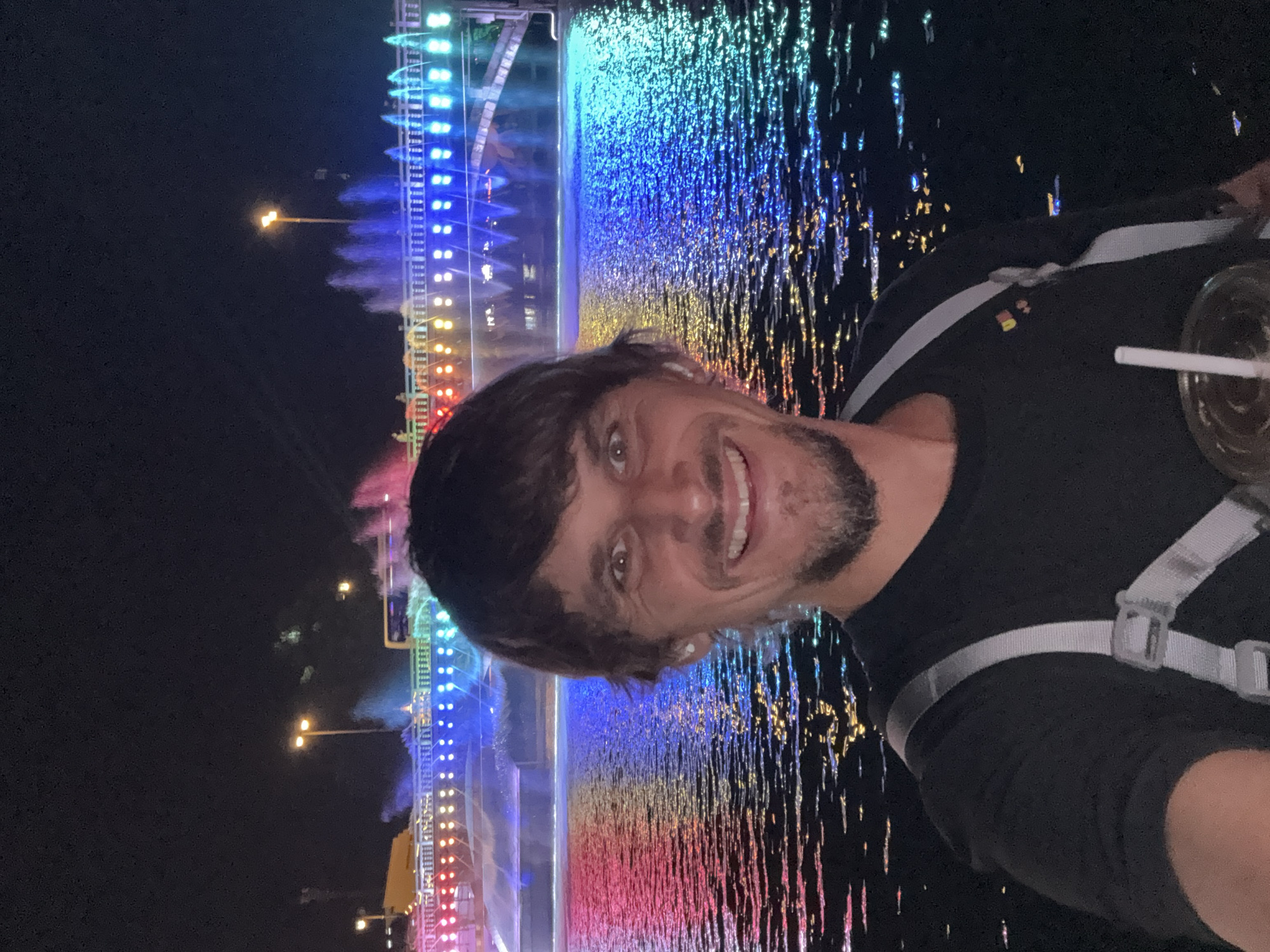 A medium shot captures a man holding a beverage in front of a colorfully lit fountain. The man has short, dark hair, a light complexion, and a light beard. He wears a black shirt with grey straps across his shoulders. The fountain displays various colors from top to bottom, including blue, green, yellow, orange, and red. The location is Wulingyuan, China.