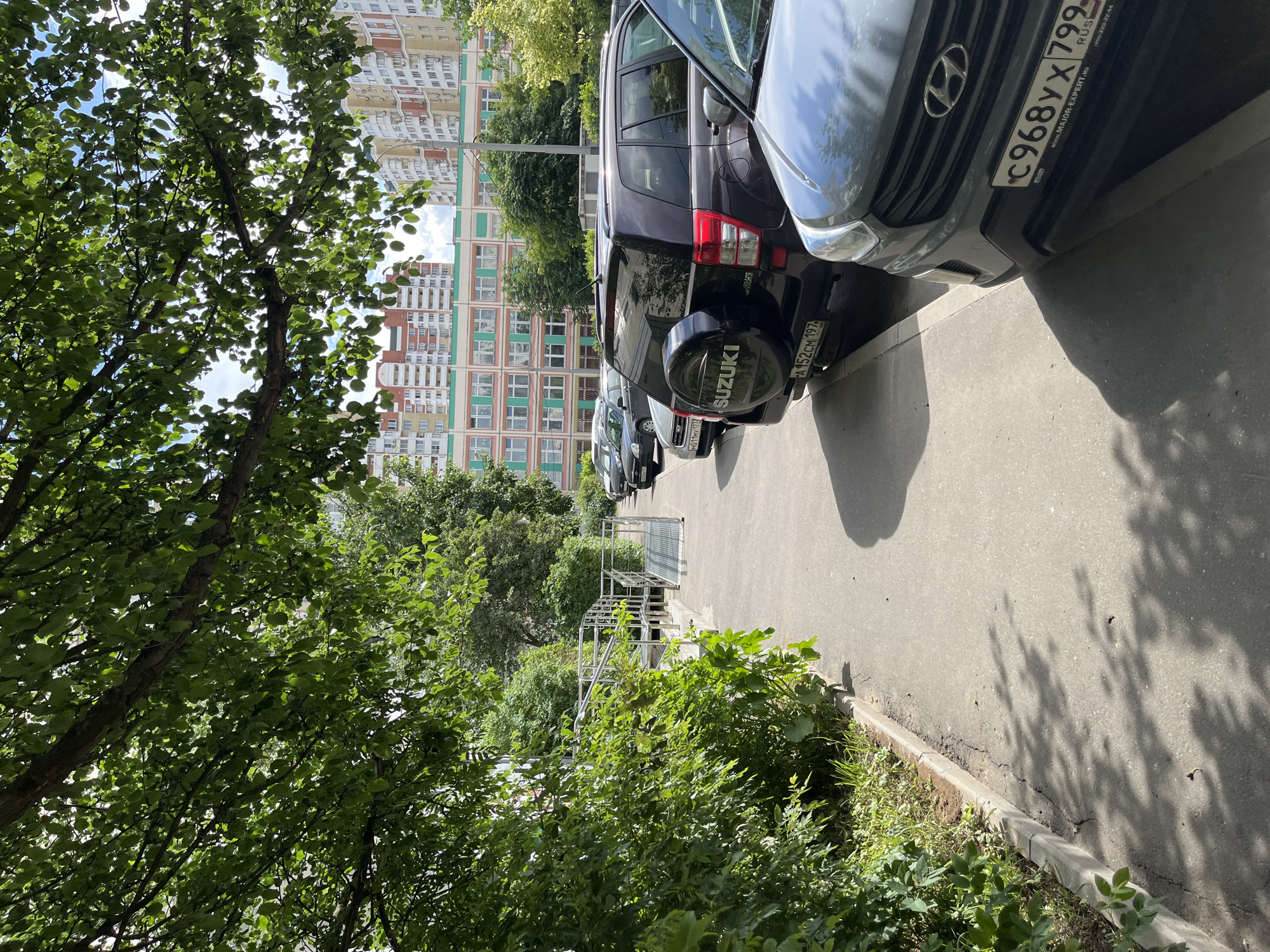 The image shows a residential street in Moscow, Russia, on a sunny day. A sidewalk runs along the left side of the frame, bordered by a lush green bush and trees. Several parked cars are visible, including a dark-colored Suzuki SUV with a spare tire mounted on the rear door, and a grey Hyundai SUV in the foreground. In the background, tall apartment buildings with red and yellow facades stand against a blue sky with scattered clouds. A wheelchair ramp leads up to a walkway between the buildings. The scene is framed by the branches and green leaves of a tree overhead, casting dappled shadows on the ground.