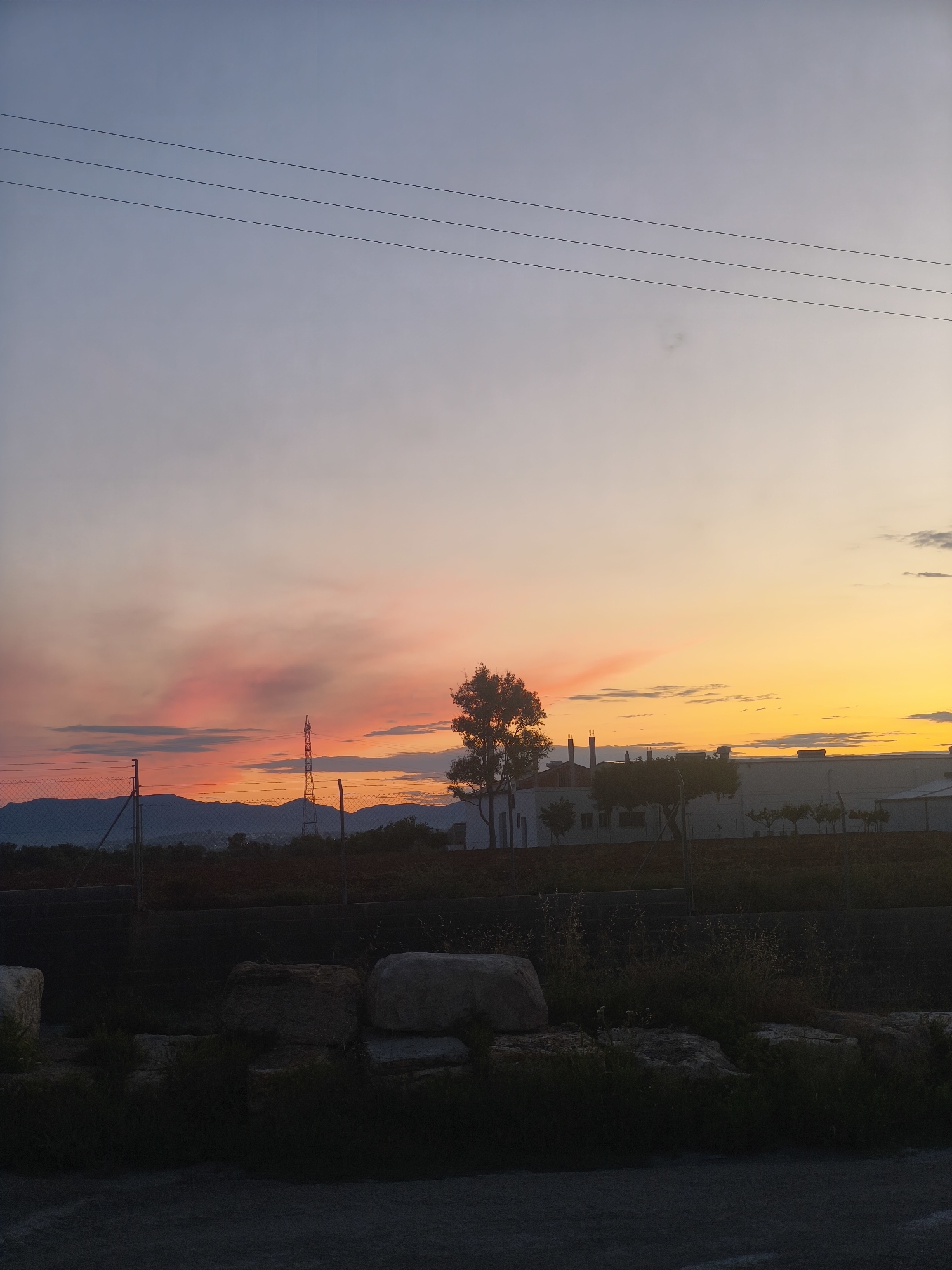 The image depicts a landscape in Riba-roja de Túria, Spain, at sunset. The sky transitions from pale blue and lavender at the top to orange and red near the horizon. A set of power lines stretches across the upper portion of the frame. Below, a chain-link fence runs horizontally across the middle ground. Beyond the fence, a building with chimneys and a line of trees is visible. In the distance, mountains can be seen. The foreground is dark, featuring rocks and vegetation, and the texture of a paved surface. An electrical tower is silhouetted against the colorful sky. The lighting is dim, typical of twilight.
