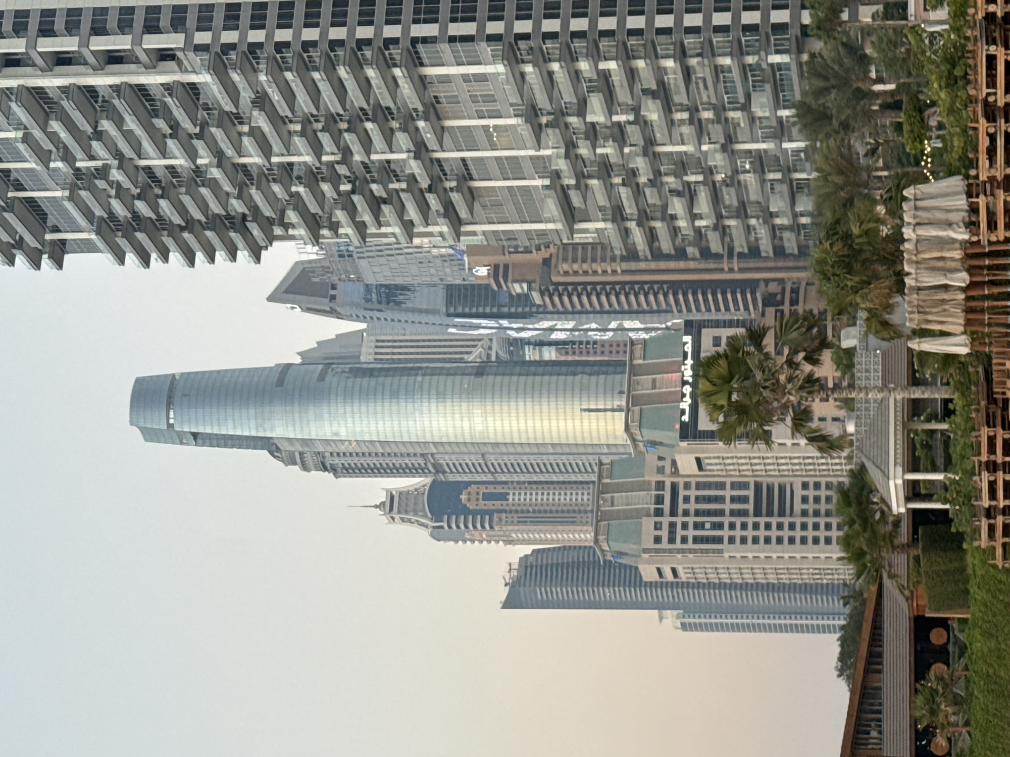 The image depicts a cityscape in Dubai, United Arab Emirates, focusing on the modern architecture of skyscrapers and residential buildings. Dominating the center is a tall, slender skyscraper with a curved, glass facade that reflects the sky. To its right, a densely packed residential building features numerous balconies with glass railings, creating a repetitive, textured pattern. In the foreground, a low-rise building with a wooden roof and outdoor seating areas, adorned with potted plants and palm trees, suggests a commercial or recreational space, possibly a restaurant or beach club. Several other skyscrapers of varying heights and architectural styles are visible in the background, contributing to the dense urban landscape. The sky is overcast and hazy, suggesting it may be early morning or late afternoon. No people are visible in the image.