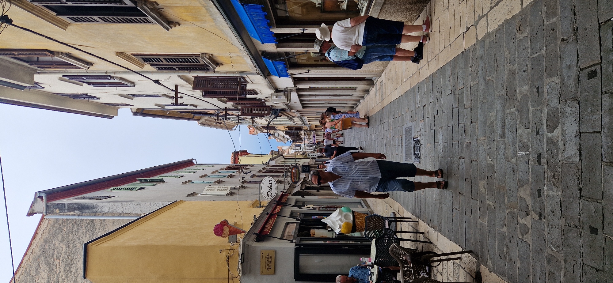 The image depicts a narrow, paved street in the old town of Koper, Slovenia. The street is lined with buildings on both sides, displaying architecture typical of Mediterranean coastal towns. The buildings have weathered facades in shades of yellow and white, with numerous windows and shutters. On the left, a building with a bright yellow facade has an oversized ice cream cone sign, indicating an ice cream shop or cafe. Further down the street, shops with awnings and display windows are visible.
Several people are present in the scene. In the foreground, a man in a striped short-sleeved shirt and denim shorts walks towards the camera. He is wearing sunglasses and sandals. To his left, a man sits at a table outside a cafe, looking down. In the middle ground and background, other individuals are seen walking, browsing shop windows, and conversing. The street is paved with stone slabs, and overhead, electrical wires crisscross the sky. The sky is overcast. The scene captures a typical day in a European old town with a mix of residents and tourists.