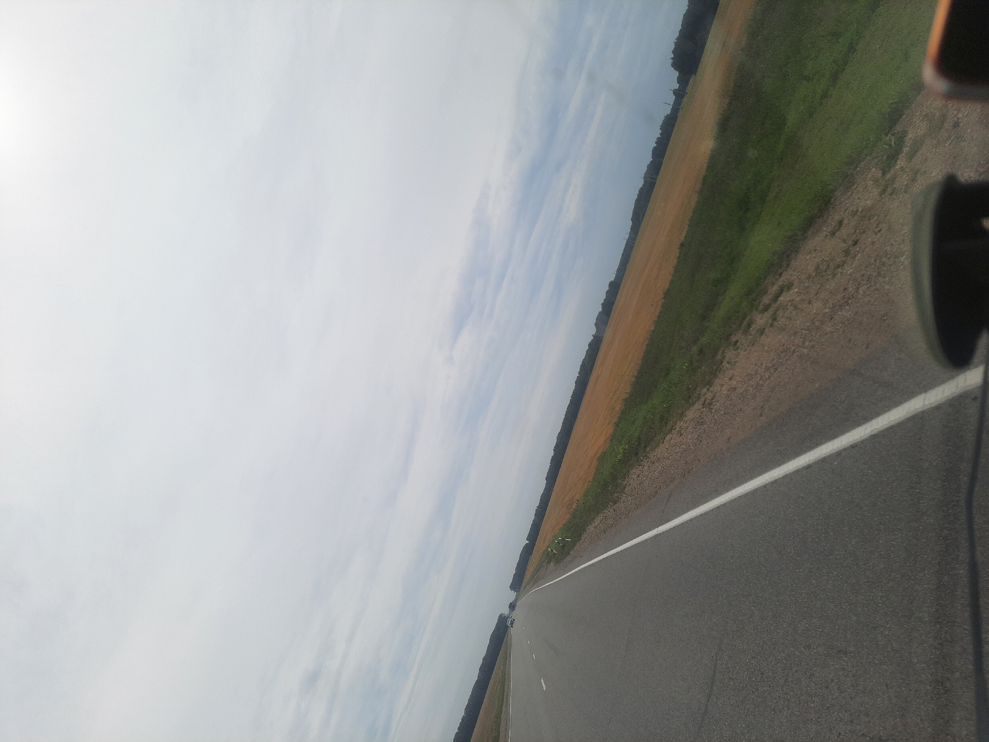 The image captures a view from a vehicle moving along a paved road in Bulanikha, Russia. The road stretches into the distance, bordered on the right by a field of golden crop and a line of trees beyond. On the left side of the road, another line of trees is visible, with a few cars driving ahead. The sky is overcast with scattered clouds. The perspective suggests the photo was taken from the driver's or passenger's seat of a car.
