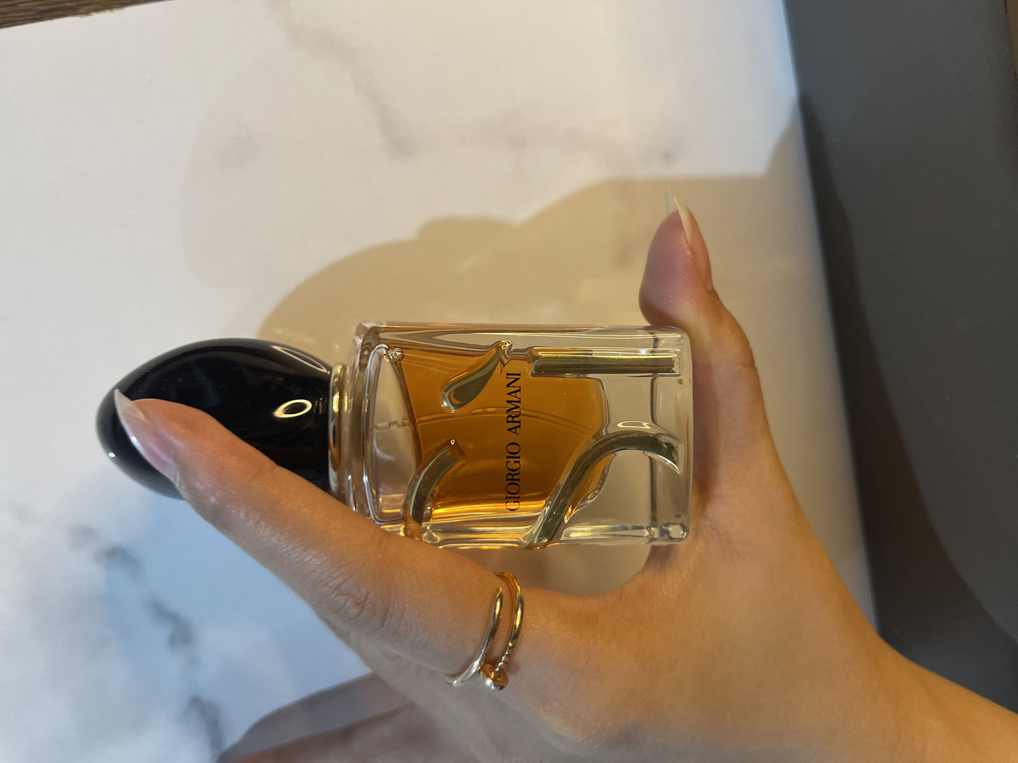 The image displays a close-up shot of a human hand holding a Giorgio Armani perfume bottle in an indoor setting. The hand, with a warm, light brown skin tone and long, neatly maintained natural nails, firmly holds the rectangular glass bottle horizontally. A delicate, gold-colored double-banded ring is worn on one of the fingers, providing a subtle accent.
The perfume bottle is transparent, allowing the rich amber-colored liquid inside to be visible. Elegant, gold-toned abstract or wavy metallic elements are integrated into the bottle's design. The bottle features a sleek, rounded black cap on its left side. Vertically printed in gold lettering on the bottle's front, the brand name "GIORGIO ARMANI" is clearly legible, with the partial word "IN" visible directly beneath it.
The background consists of muted tones. To the left, a light-colored surface with a soft, mottled texture is visible, possibly part of a book or a table. To the right, a darker, solid gray or black surface provides a contrasting backdrop. The lighting appears soft and even, suggesting an indoor environment. The image is a product-focused shot, highlighting the perfume bottle and the hand holding it, captured in Surabaya, Indonesia.
