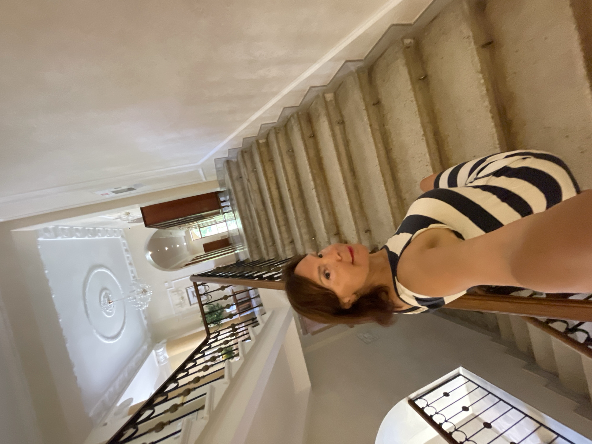 In Gagra, Abkhazia, a woman with brown hair and red lipstick, wearing a striped dress, takes a selfie on a grand staircase. The staircase features stone steps and ornate railings. The background reveals a high-ceilinged hallway with intricate plasterwork on the ceiling, a chandelier, and doorways leading to other parts of the building. The overall impression is of an elegant, classical interior.