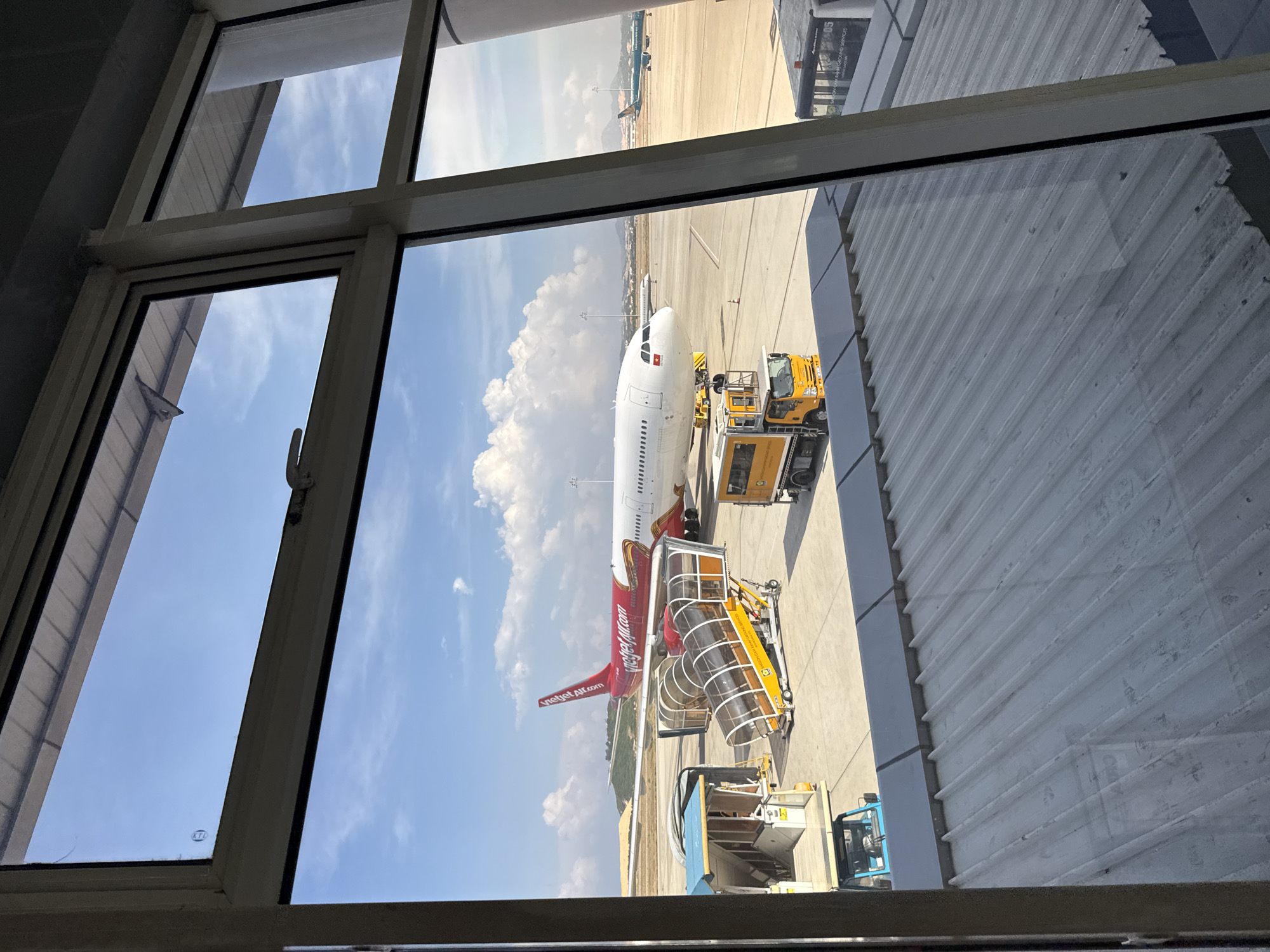 A view from a window in Bac Cam Ranh Ward, Vietnam, shows an airplane, a Vietjet Air aircraft, on the tarmac. The plane is positioned alongside boarding stairs and service vehicles. The sky above is partly cloudy. The view is partially obstructed by the window frame and adjacent building structures.