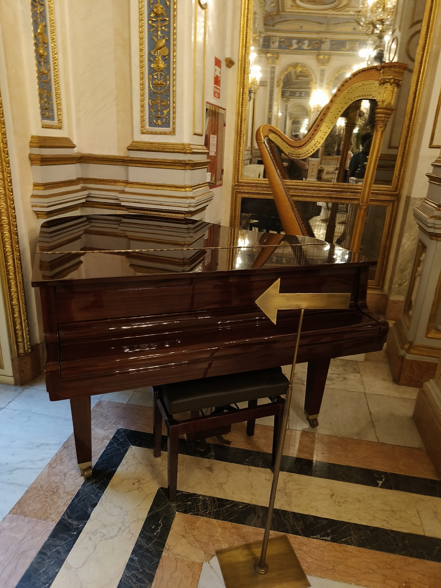 The image shows a dark brown, polished grand piano with a black seat in front of it. The piano is situated on a marble floor with geometric patterns. To the left of the piano, there is a harp reflected in a large ornate mirror. The room appears to be decorated with gold accents and molding. A brass arrow-shaped sign points to the left, placed in front of the piano. In the background, a chandelier is visible in the reflection. The overall setting suggests a formal or ornate interior, possibly a music hall or a grand room. The location is specified as the Palau de la Música in Valencia, Spain.