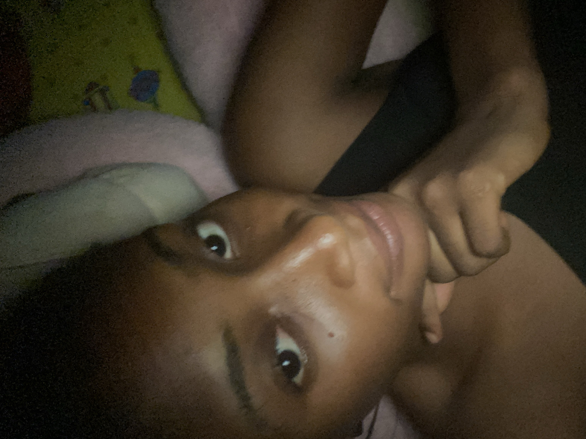 A dark-skinned individual is lying down, with their head resting on a pillow. They are looking directly at the camera with a neutral expression. The lighting in the room is dim, creating a shadowy atmosphere. The background consists of a blurred green and pink patterned surface, possibly a blanket or a rug. The image appears to have been taken indoors, likely in a bedroom. There are no visible landmarks or contextual clues to indicate the specific location within Kubwa, Nigeria. The subject is wearing a dark colored top.