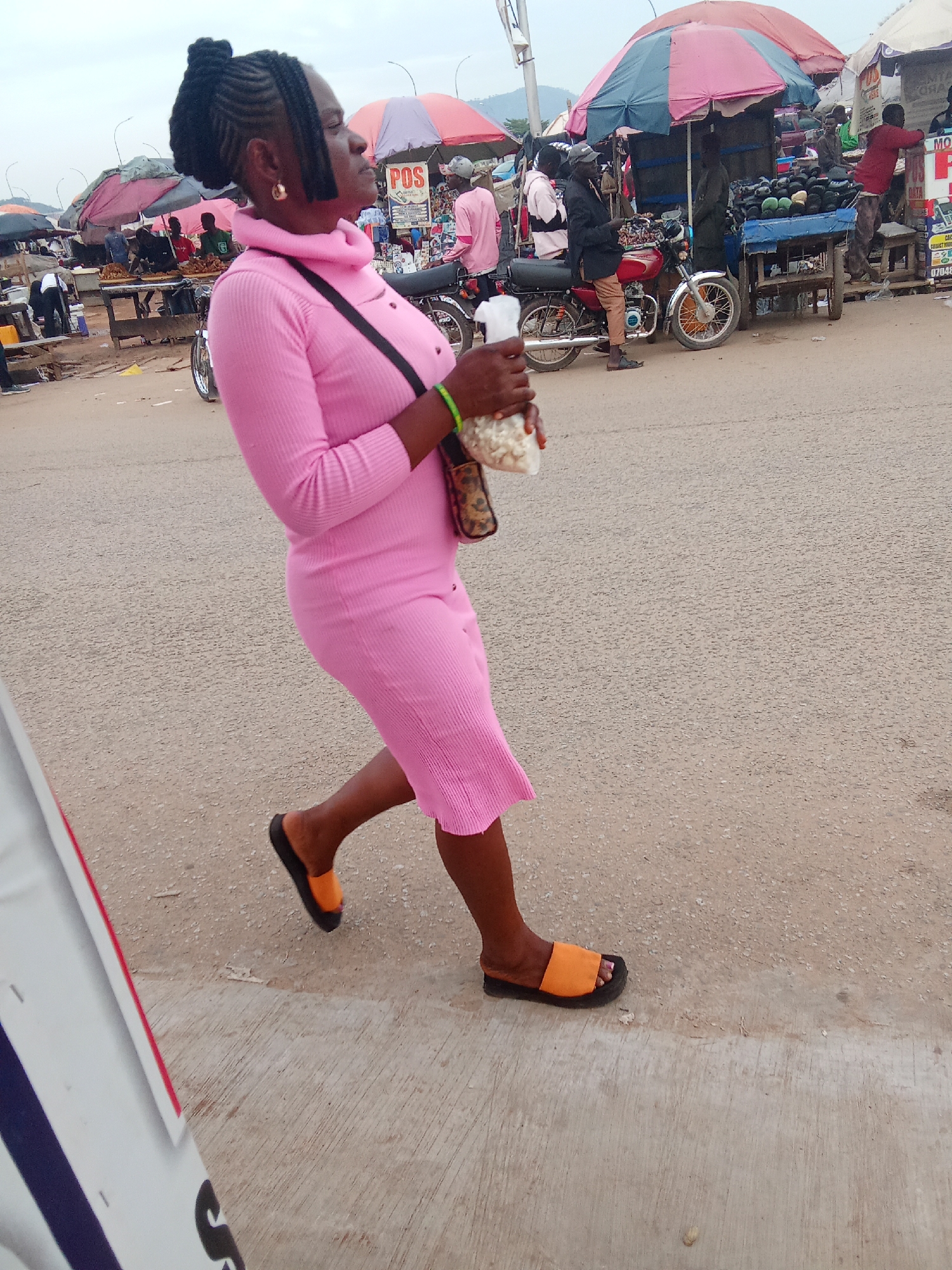 In Kubwa, Nigeria, a woman in a pink, form-fitting, long-sleeved dress with a high neckline walks across a paved surface. Her hair is styled in braids with short, blunt bangs. She wears large gold hoop earrings and orange and black slide sandals. A leopard-print purse hangs across her body, and she holds a clear plastic bag filled with what appears to be popcorn in her right hand. The background depicts an outdoor market scene with various stalls and umbrellas, a motorcycle parked nearby, and other people visible among the vendors and customers. The overall atmosphere suggests a bustling marketplace.