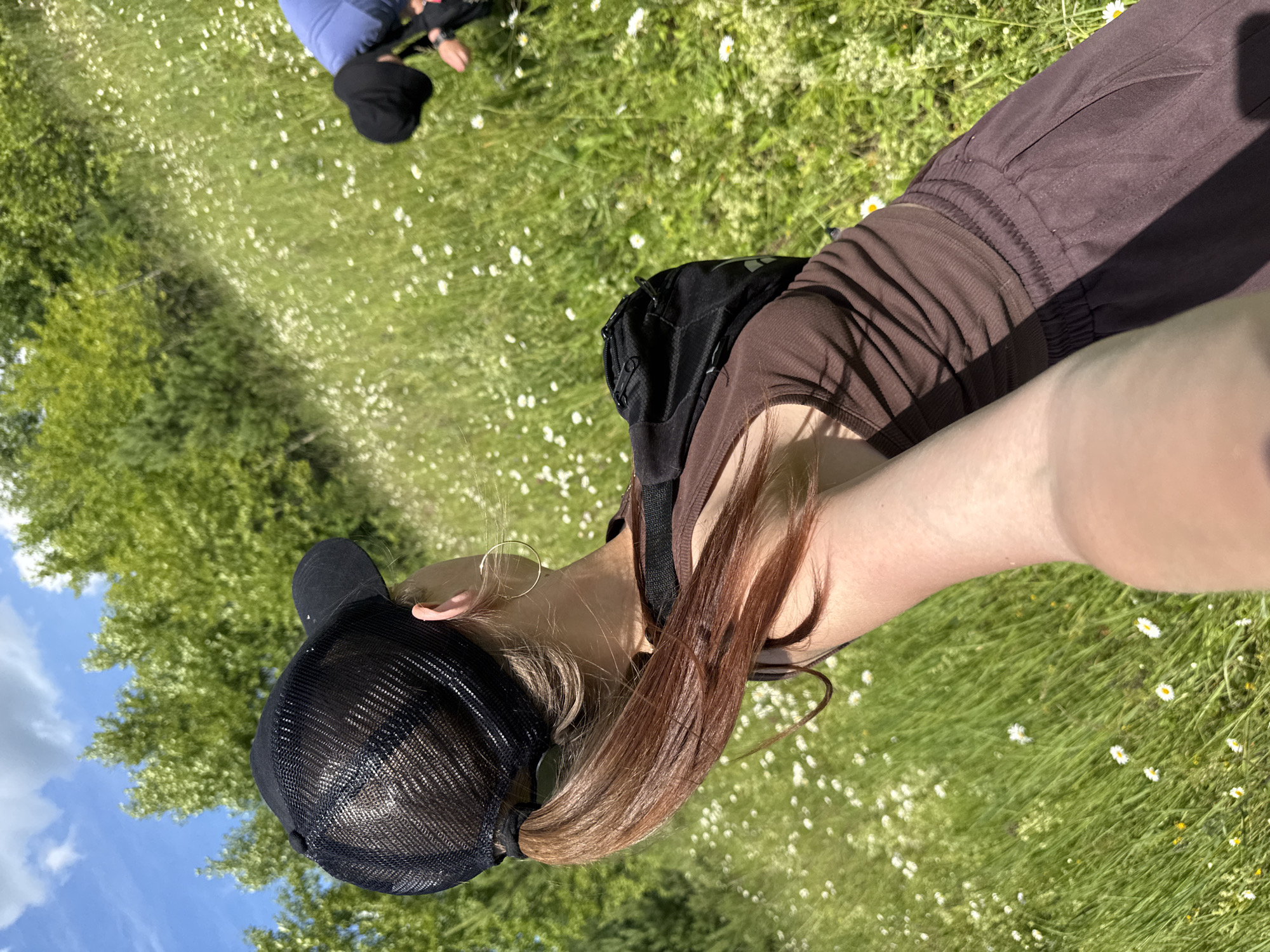 A woman wearing a black mesh baseball cap and a brown tank top stands in a grassy field with small white flowers. She has her hair in a ponytail and is wearing a large hoop earring. Her right arm is extended towards the camera, as if taking a selfie. In the background, another person in a blue shirt and black hat is partially visible, bending down to examine the flowers. The field is surrounded by green trees, and the sky is blue with scattered white clouds. The image appears to be taken outdoors on a sunny day, possibly in a park or rural area near the village of Garevaya, Russia.