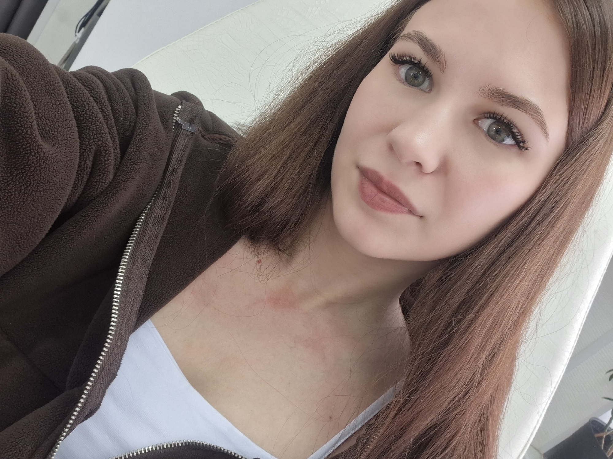A young woman is captured in a close-up selfie, looking directly at the camera with a calm expression and a soft, slight smile. She has long, straight, light to medium brown hair that falls over her shoulders. Her eyes are a prominent feature, appearing large and a light green or hazel color, complemented by full, dark eyelashes, possibly extensions. Her eyebrows are well-defined and neatly groomed. She is wearing light makeup, including a subtle pinkish-brown lip color.
She is dressed in a dark brown, textured, fleece-like zippered hoodie or jacket, worn over a white tank top or undershirt. The silver-toned zipper of her jacket is visible down her front. On her neck and upper chest, several faint reddish marks or blotches are visible, along with a small mole.
The setting appears to be indoors, with a plain white or light-colored surface, possibly a wall or the back of a chair, visible behind her. A dark object, which might be a plant in a pot, is partially visible in the bottom right corner of the background. The lighting suggests an indoor, possibly daylight, setting. No discernible text is visible in the image. The photo provides a close personal view, likely taken in Chaikovsky, Russia.