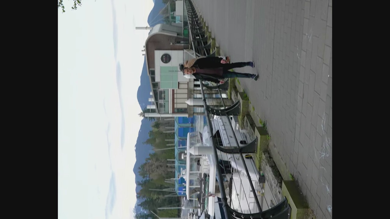 A man is walking on a paved path next to a marina. He is wearing a dark jacket, a maroon shirt, and jeans. In the background, there are boats docked in the water and a modern building with large windows. Mountains are visible in the distance, covered in trees and snow. The sky is overcast. The scene appears to be in Vancouver, Canada, likely at Coal Harbour Marina, given the presence of the distinctive building and the mountain backdrop.