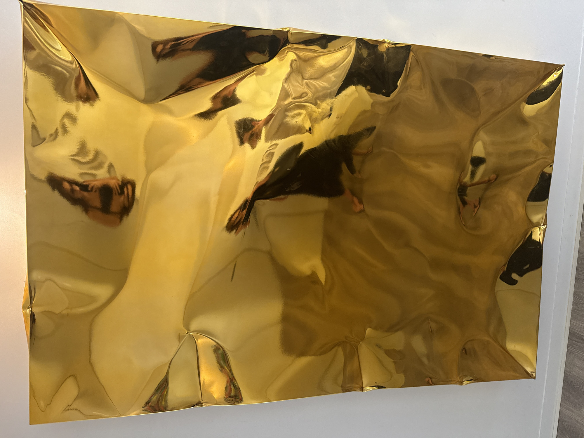 The image shows a large, crumpled, gold-colored sheet of material mounted on a white wall. The material is highly reflective, mirroring the surroundings and creating distorted images. In the upper left quadrant, a reflection shows a person, possibly a woman, holding a phone and taking a selfie. The person has dark hair pulled up. In other areas of the reflection, partial figures and objects can be seen, suggesting the sheet is in a room with other elements. The bottom edge of the gold sheet rests on a white baseboard, and the floor appears to be a light-colored wood. The lighting comes from above and to the left, casting highlights on the metallic surface. There is no indication of Grândola, Portugal in the image itself.