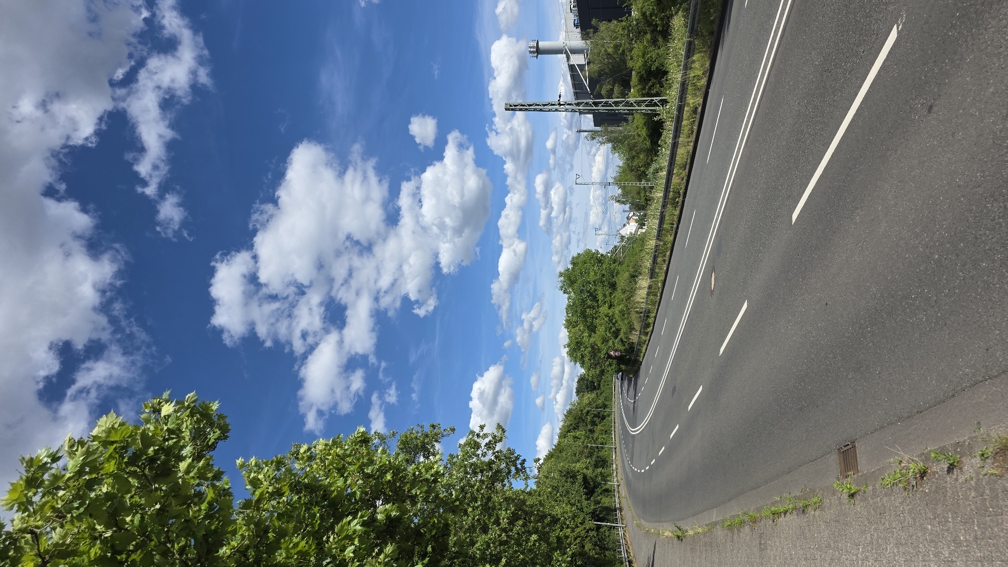 A winding asphalt road with two lanes in each direction is pictured under a bright blue sky dotted with white cumulus clouds. To the left, lush green trees line the roadside, with a concrete sidewalk at the very edge. The road curves to the right, leading into the distance where a few buildings and industrial structures, including a tall smokestack, are visible amidst more greenery. Overhead power lines are also present in the background. The scene appears to be on the outskirts of a city, with a clear and sunny atmosphere.