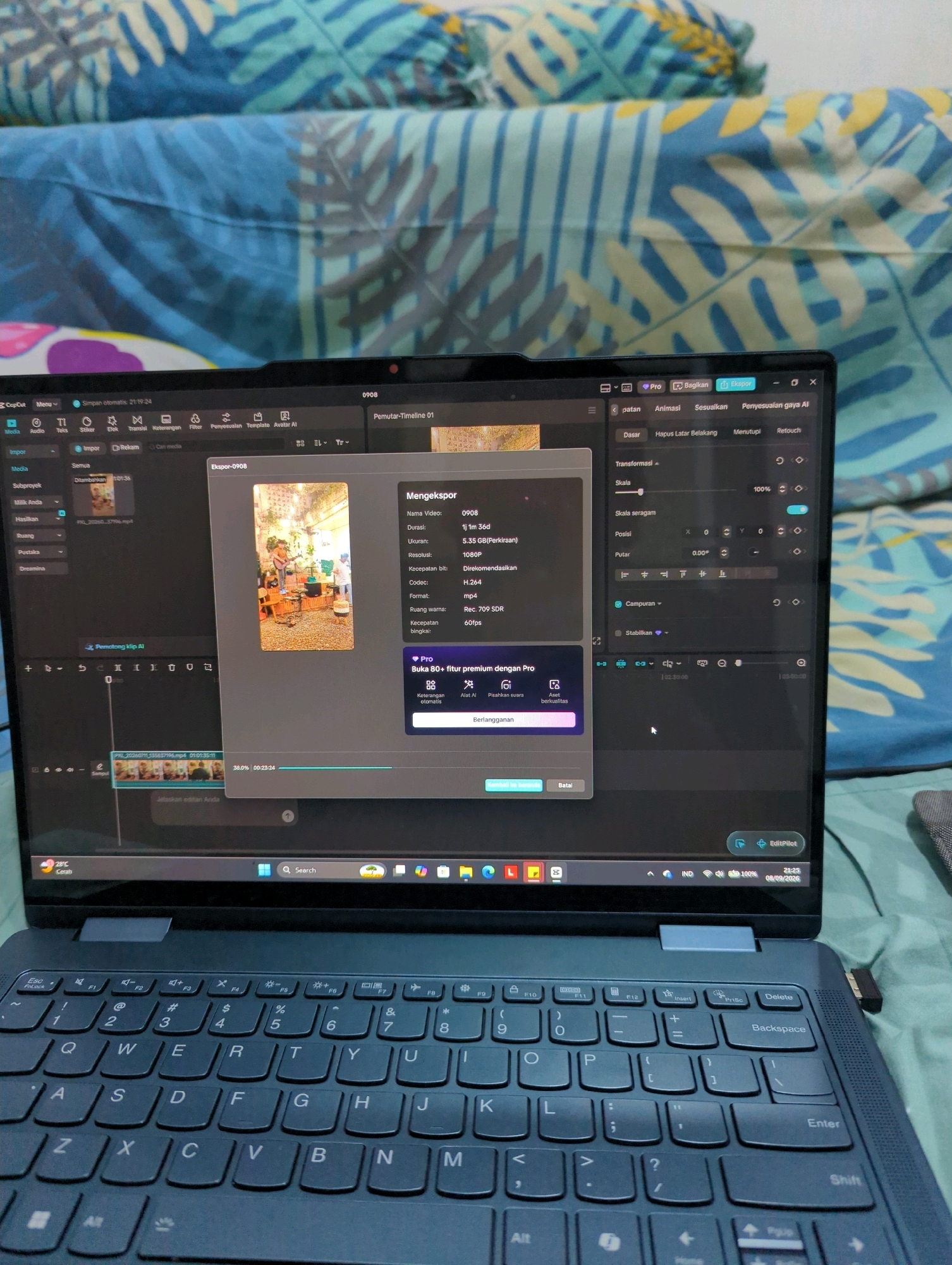 This is a close-up, eye-level shot of a laptop screen displaying video editing software, specifically CapCut, with a video export window open. The laptop is open and placed on a surface covered by a blue and green patterned fabric, likely a bedspread or blanket, which is characteristic of a home environment in Depok, Indonesia. The laptop's keyboard is partially visible in the foreground, showing a standard QWERTY layout. The screen shows a preview of a video clip with a person playing a guitar in what appears to be an indoor cafe setting. The export window details the video's properties, including its name "0908", duration "1j 1m 36d", resolution "1080P", and codec "H.264". The overall scene suggests a person is in the process of exporting or reviewing a video project on their laptop.