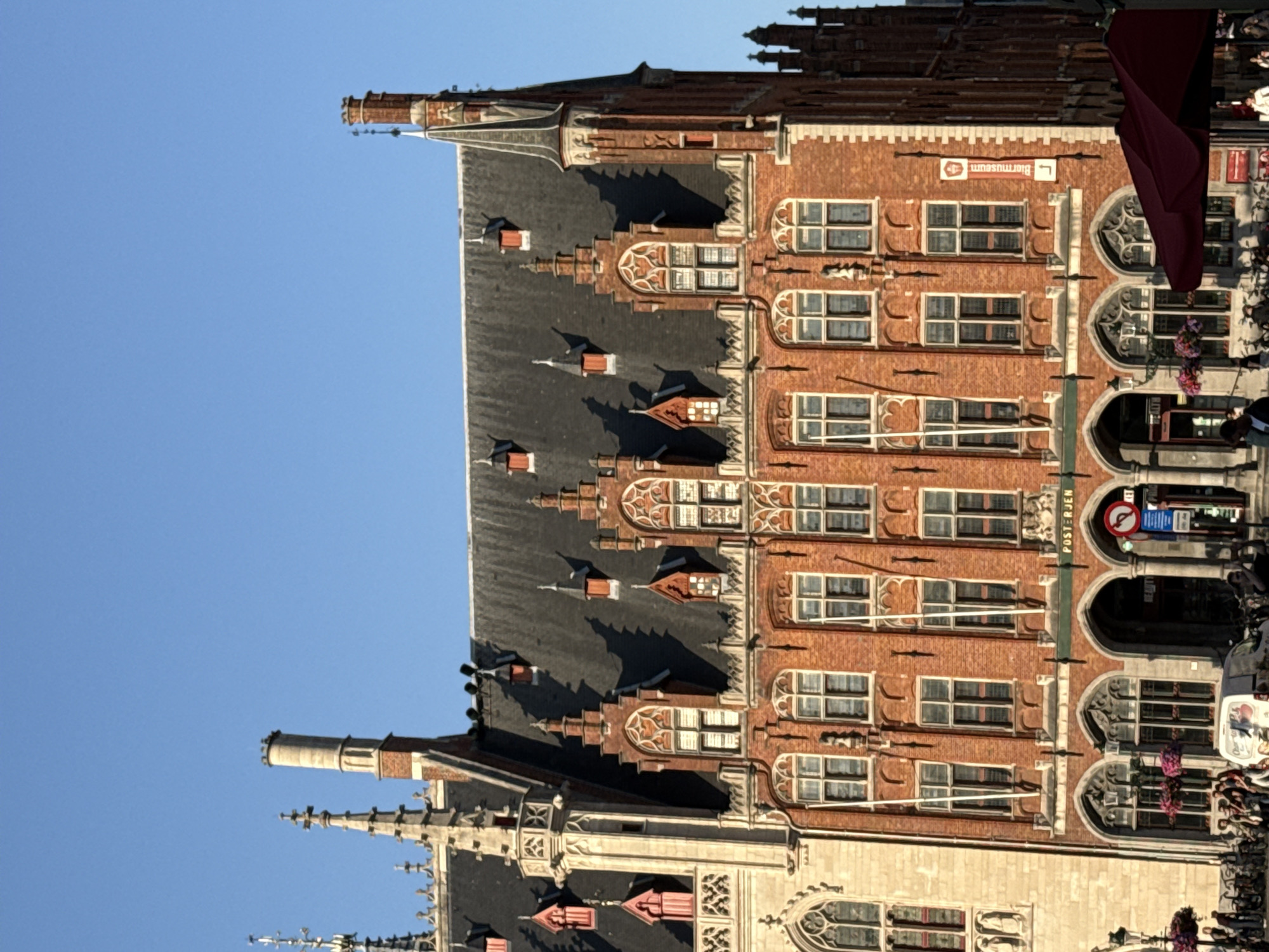 The image depicts the exterior of the historic Post Office building in Bruges, Belgium, under a clear blue sky. The building is constructed of red brick with intricate Gothic architectural details, including pointed arched windows, ornate gables, and a steep, dark-tiled roof. Several chimneys and spires rise from the roofline. The facade features multiple levels of windows, some with decorative tracery. A sign with the word "POSTJEB" is visible above an arched entryway. In the foreground, several people are walking or standing, and bicycles are parked. A red umbrella is partially visible on the right side of the frame. The overall scene suggests a vibrant urban environment in a historic European city.
