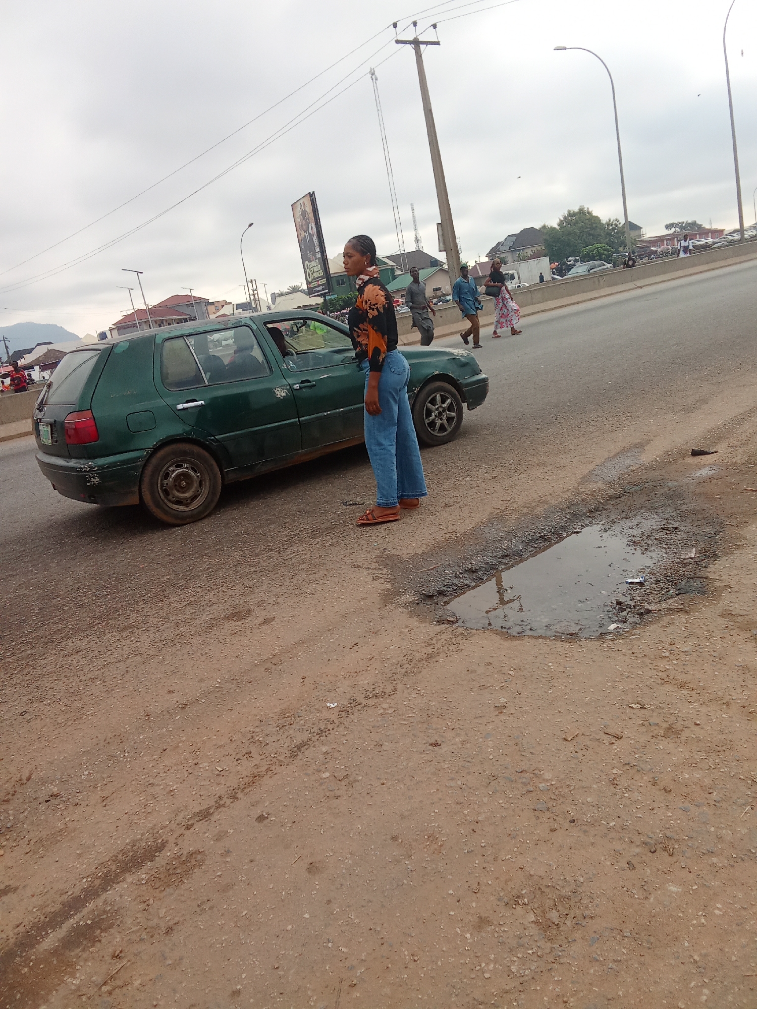 A full shot shows a green Volkswagen Golf Mk3 hatchback parked on a dirt road adjacent to a paved road in Kagini, Nigeria. The car is facing left, with its driver's side door ajar. A young woman wearing a black and orange patterned shirt and blue jeans stands next to the driver's side of the car, looking to her right. A puddle of water and debris are present on the dirt road in the foreground. In the background, several people are walking on the paved road, and a billboard is visible on the right side of the street. The sky is overcast, and the general lighting is dim.