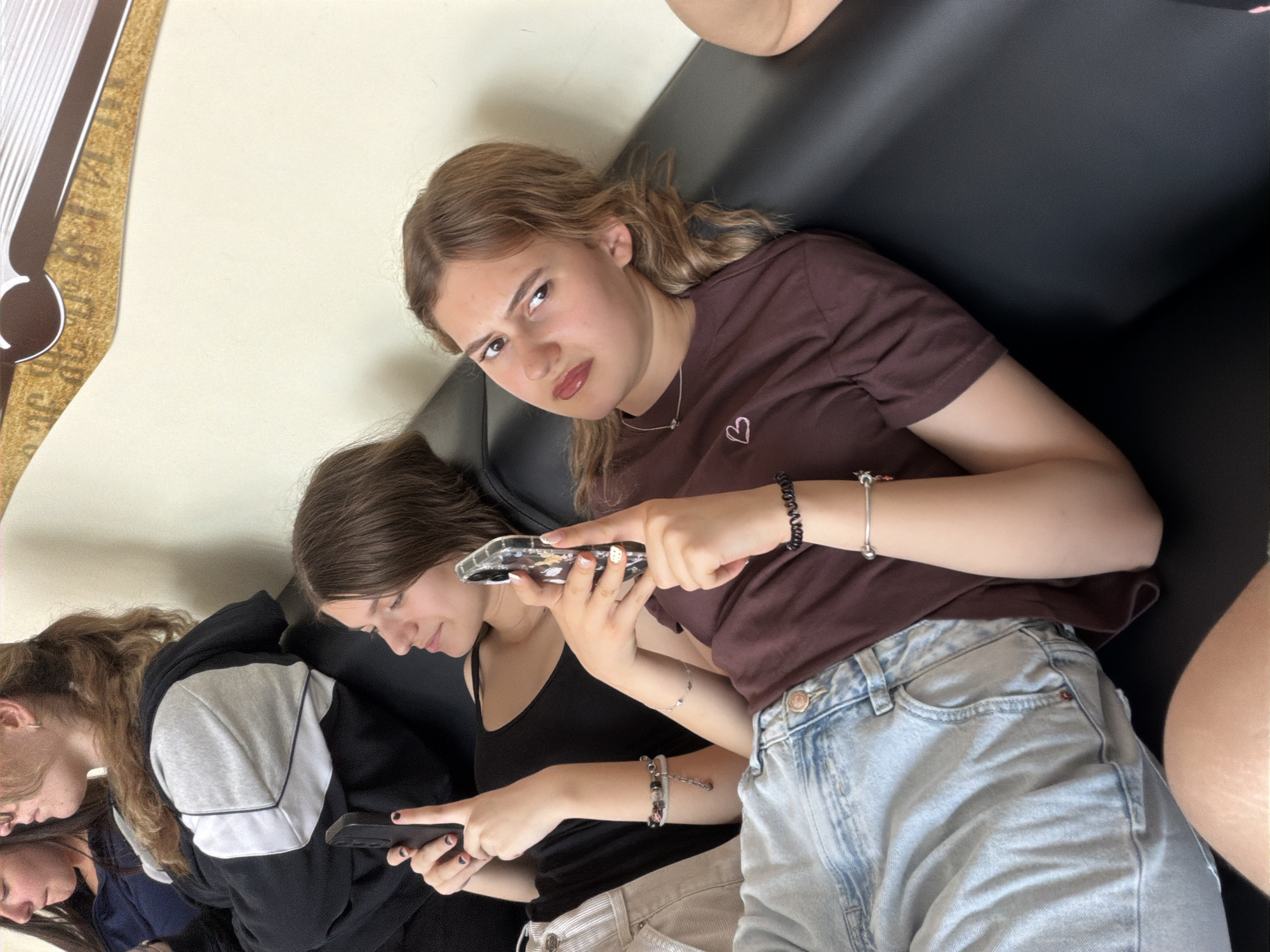 Three young women are seated indoors on a black sofa. The image is taken from a slightly elevated, angled perspective.
The central figure, a young woman with light brown, wavy hair, is looking directly at the camera with a skeptical or annoyed expression, her eyebrows furrowed and lips slightly pursed. She is wearing a brown t-shirt with a small pink heart embroidered on the chest, and light blue denim jeans. She holds a smartphone in her left hand, pointing it with her right index finger towards the woman seated below her. She also wears a silver necklace and multiple bracelets on her wrists.
Below her, another young woman with dark brown hair is visible from the chest up. She is engrossed in her own smartphone, looking down at the screen. She wears a black tank top and light-colored pants, along with several bracelets.
On the far left, a third young woman is partially visible. She has light brown hair and is wearing a black and grey hoodie, also looking downwards, presumably at a device or activity not fully captured.
The background consists of a plain light-colored wall. To the far left, a decorative element, possibly a frame or wall art, features brown and white stripes with some indistinct dark patterns or text on a light brown background. The overall atmosphere suggests a casual, indoor setting, where the individuals are relaxing and engaged with their mobile phones. No specific location cues are visible beyond the provided context of Panagyurishte, Bulgaria.