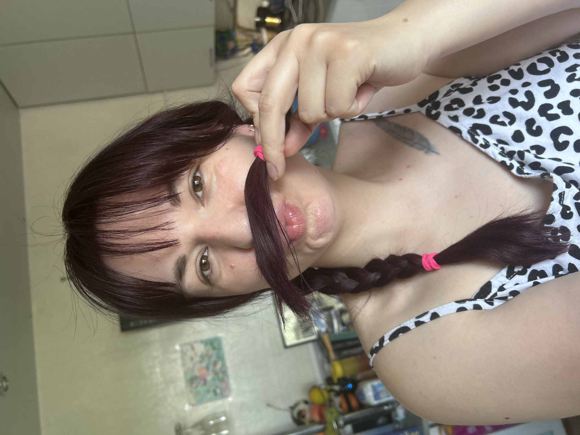 The image, captured in a domestic setting within Foki, Russia, presents a Caucasian female from the chest up, centered and looking directly at the camera. She has dark auburn hair with straight bangs, styled with a single braid extending over her left shoulder, secured by a bright pink elastic band. She is holding a portion of her braided hair across her upper lip, mimicking a mustache, while her lips are puckered in a "kissing" or "fish face" expression. Her brown eyes are open, and minor skin blemishes are visible on her face. She is wearing a white camisole with a black leopard print pattern. A grayscale tattoo resembling a feather is visible on her upper left chest/shoulder area. The background features light-colored walls. To the left, a blurry colorful picture hangs on the wall, and various household items including glassware, figurines, and bottles are visible on shelves or a counter. To the right, blurred light-colored kitchen cabinets are partially discernable.