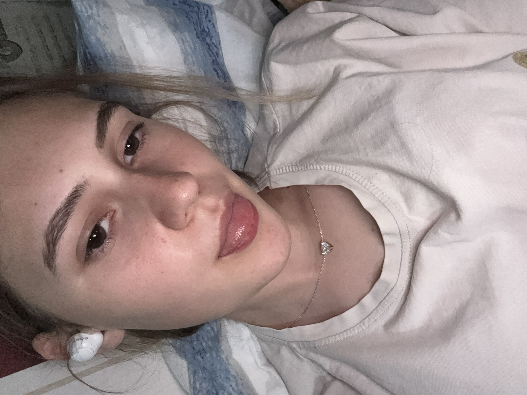 A young female with light brown hair is lying down and sticking her tongue out. She is wearing white earbuds in her left ear and a light-colored t-shirt with a heart-shaped pendant necklace. She is in a room with blue and white striped bedding in the background. There is no information or visual indication of the location being in Panagyurishte, Bulgaria.